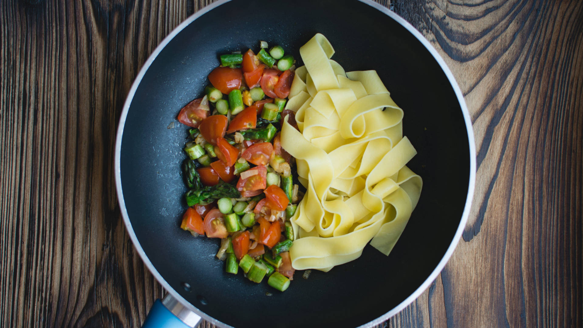 Pasta 桌布 壁紙 — 新鮮義大利麵和色彩繽紛的炒蔬菜並排放在黑色煎鍋中，置於鄉村風格的木質表面上。