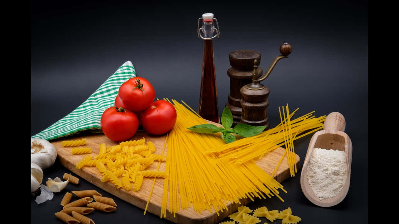 720p Pasta 桌布 壁紙 — 新鮮義大利麵材料如番茄、大蒜、羅勒和麵粉，排列在木板上，背景深色。