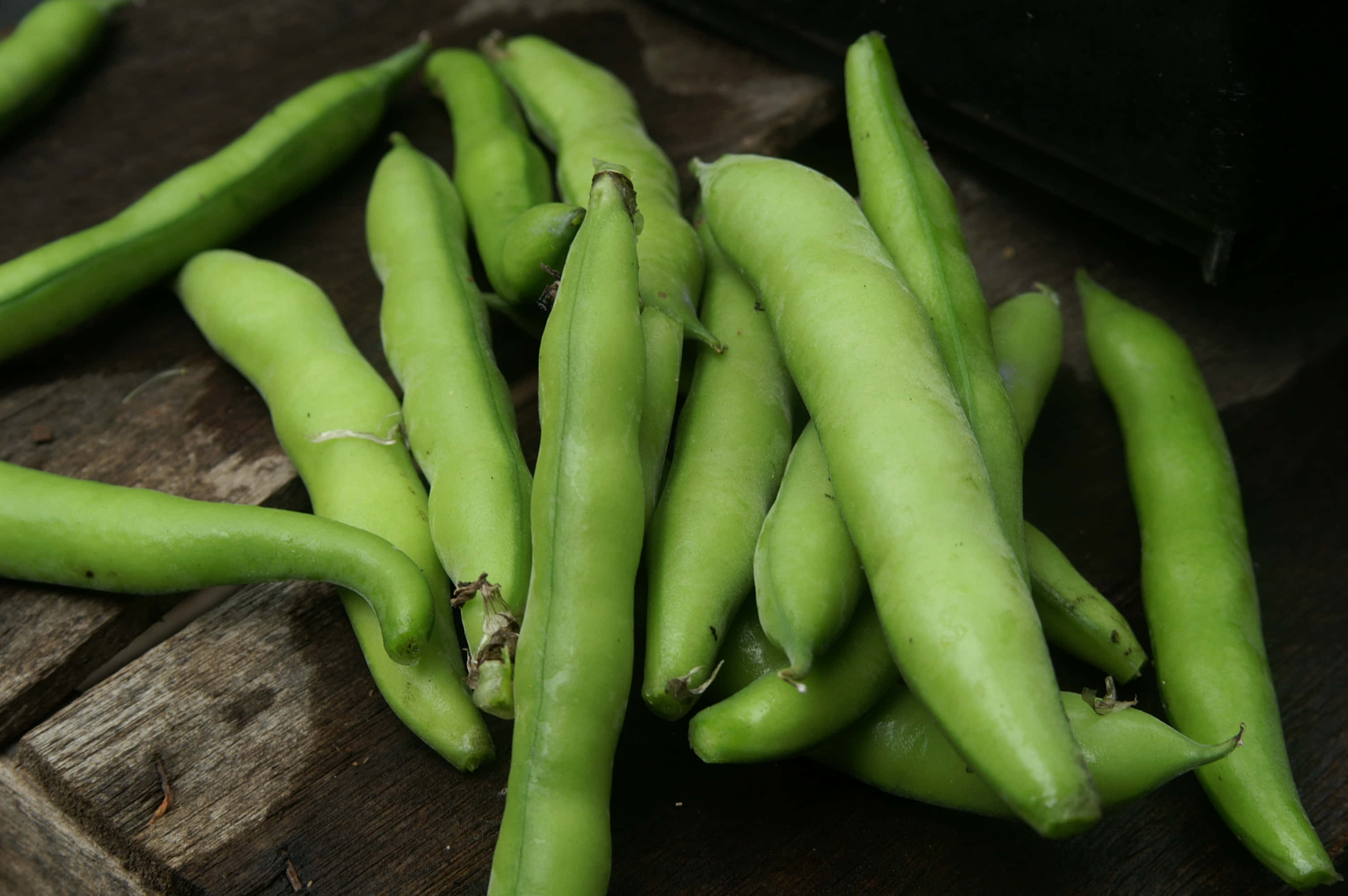 beans 桌布 壁紙 — 新鮮翠綠的蠶豆莢堆放在經風化的木質表面上，柔和自然光線照射。
