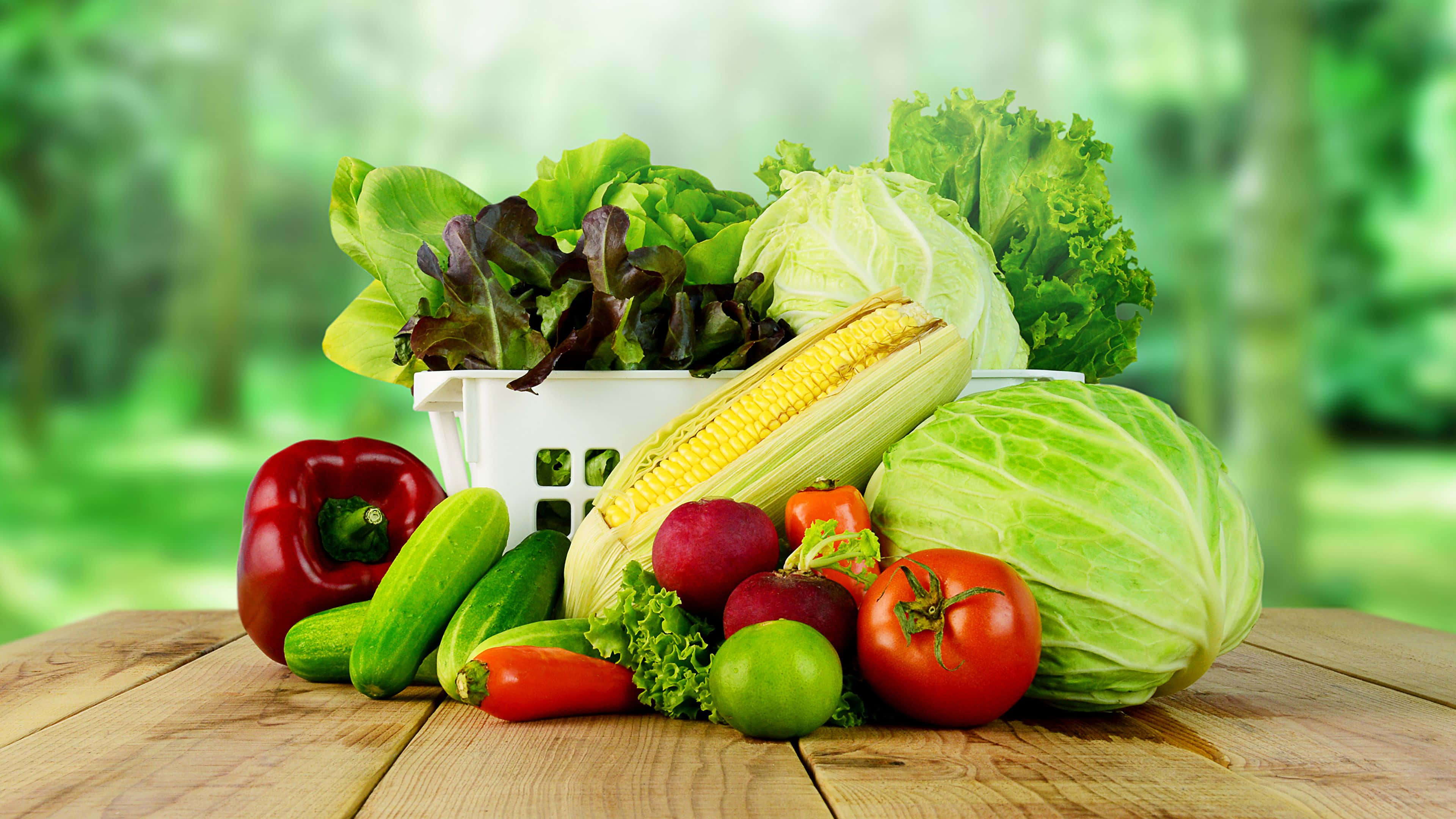 Vegetables 桌布 壁紙 — 新鮮色彩繽紛的蔬菜，如番茄、玉米和生菜，擺放在木桌上，背景為模糊的綠色。