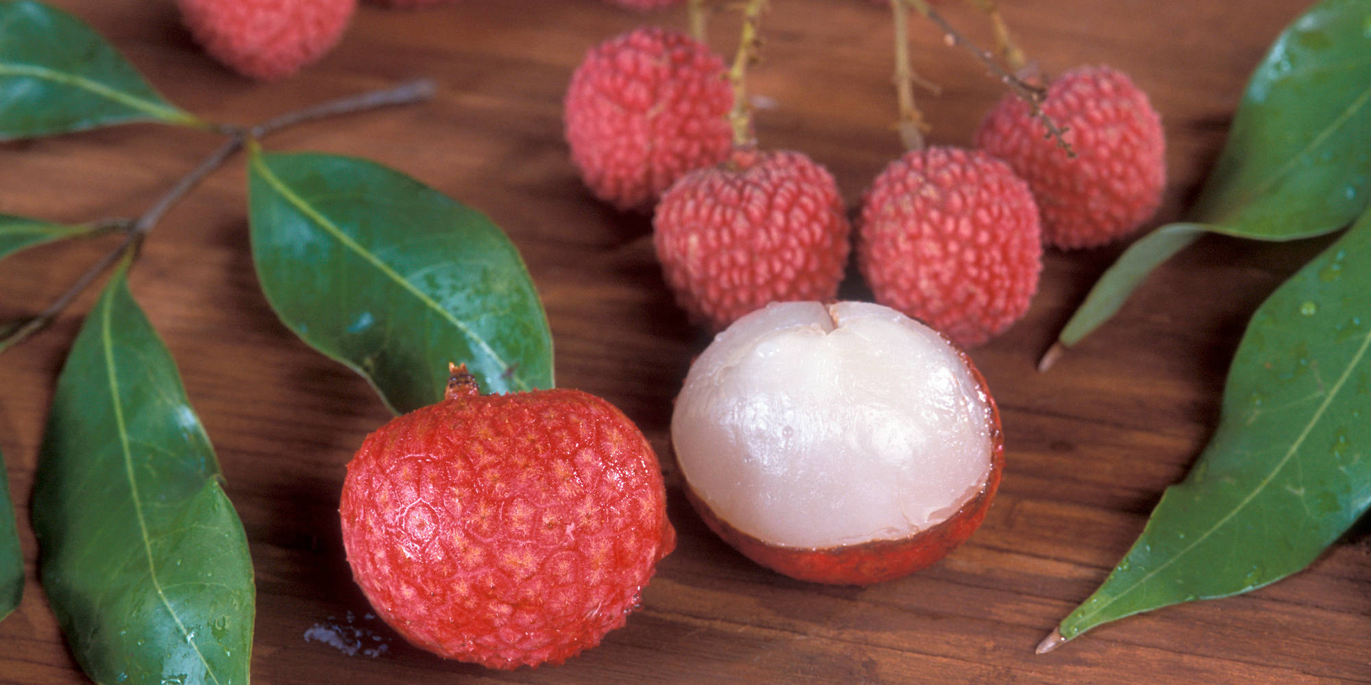 Litchi 桌布 壁紙 — 新鮮荔枝帶有光澤的紅色外皮和半透明的白色果肉，放在木質表面上，旁邊有綠葉。