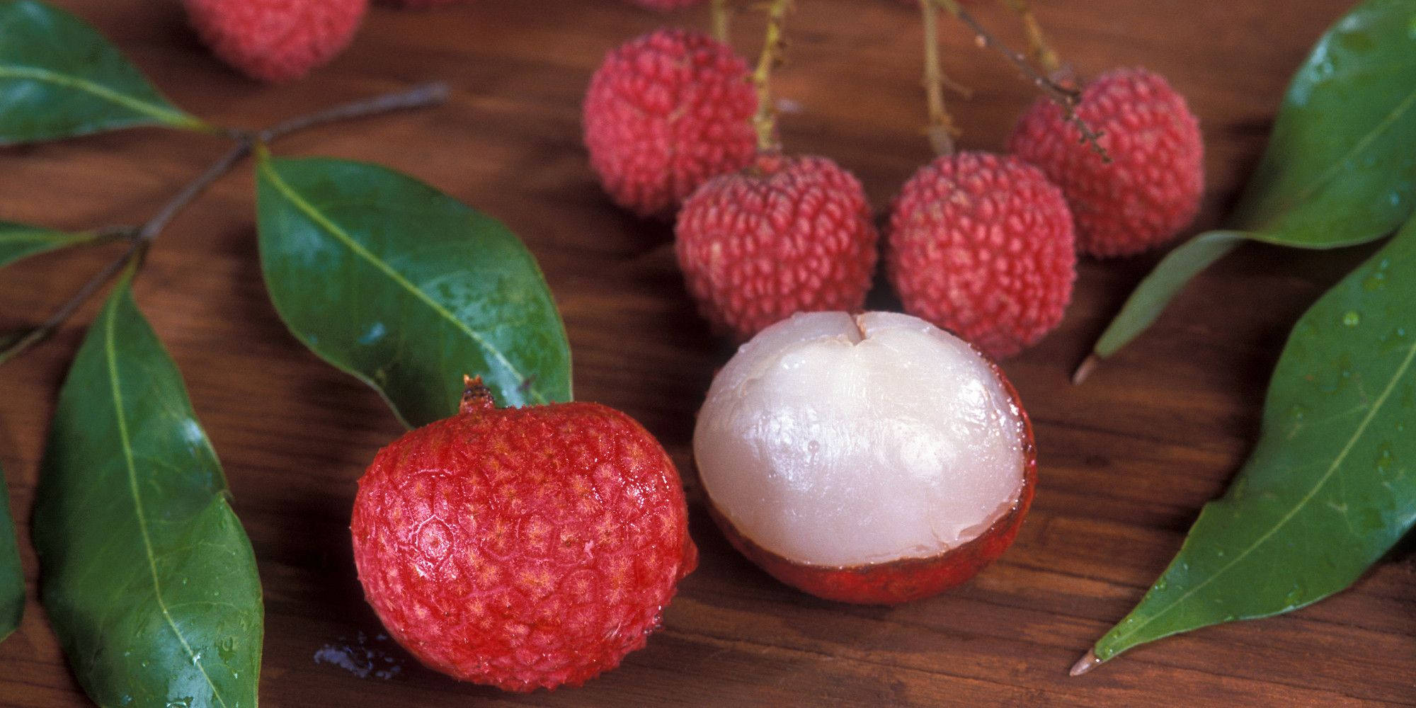Fruits 桌布 壁紙 — 新鮮荔枝帶有光澤的紅色外皮和半透明的白色果肉，放在木質表面上，旁邊有綠葉。