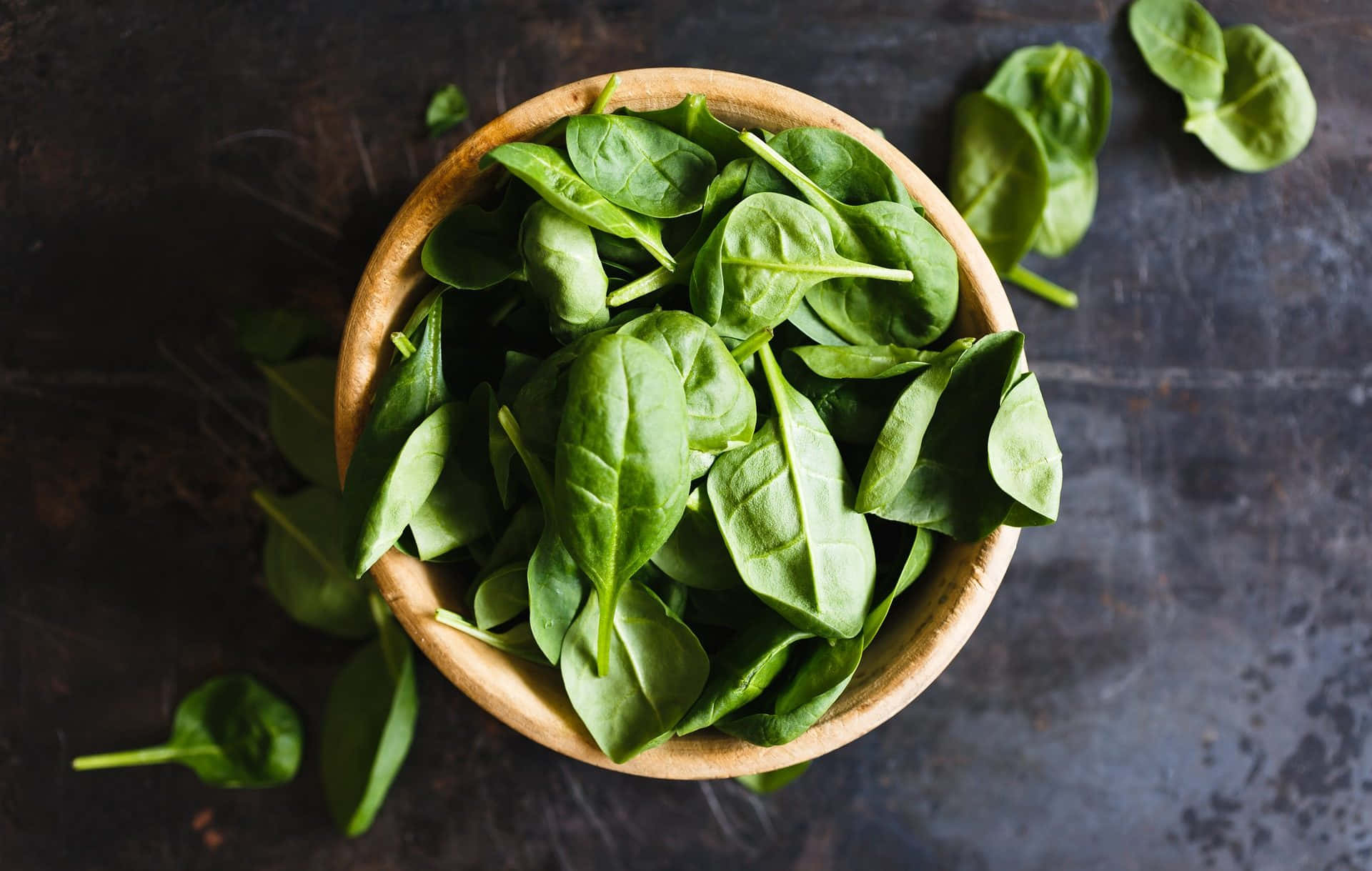 Vegetables 桌布 壁紙 — 新鮮菠菜葉從木碗中溢出，落在深色紋理表面上，俯拍視角。