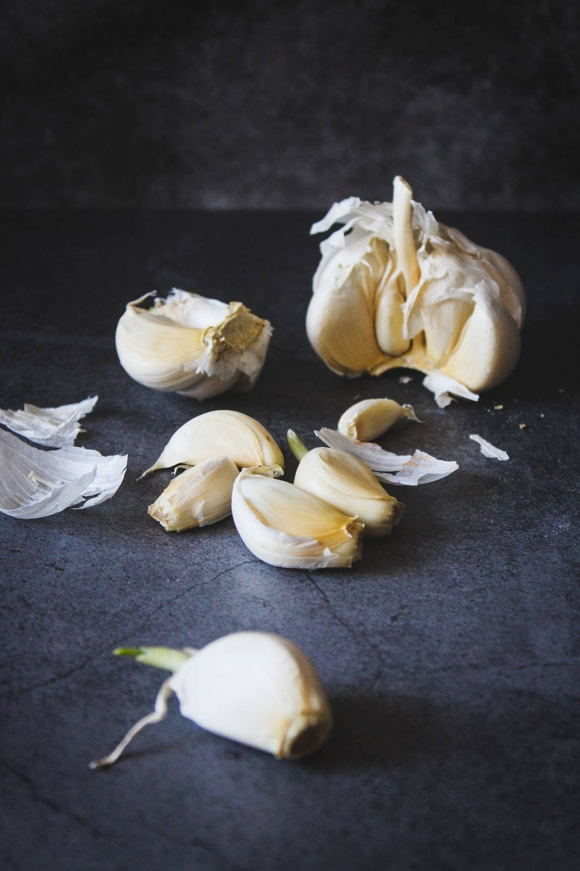 Garlic 桌布 壁紙 — 新鮮蒜瓣和蒜頭散落在深色紋理表面上，光線柔和。