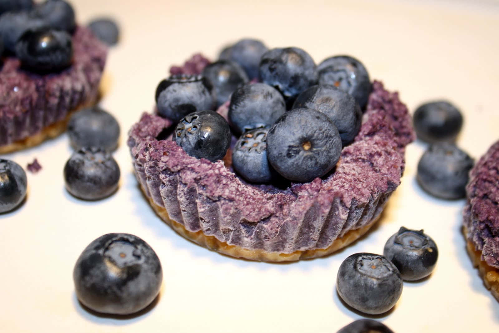 Blueberries Tart 桌布 壁紙 — 新鮮藍莓塔的微距特寫，帶有紫色餡料和散落的莓果，置於白色表面上。