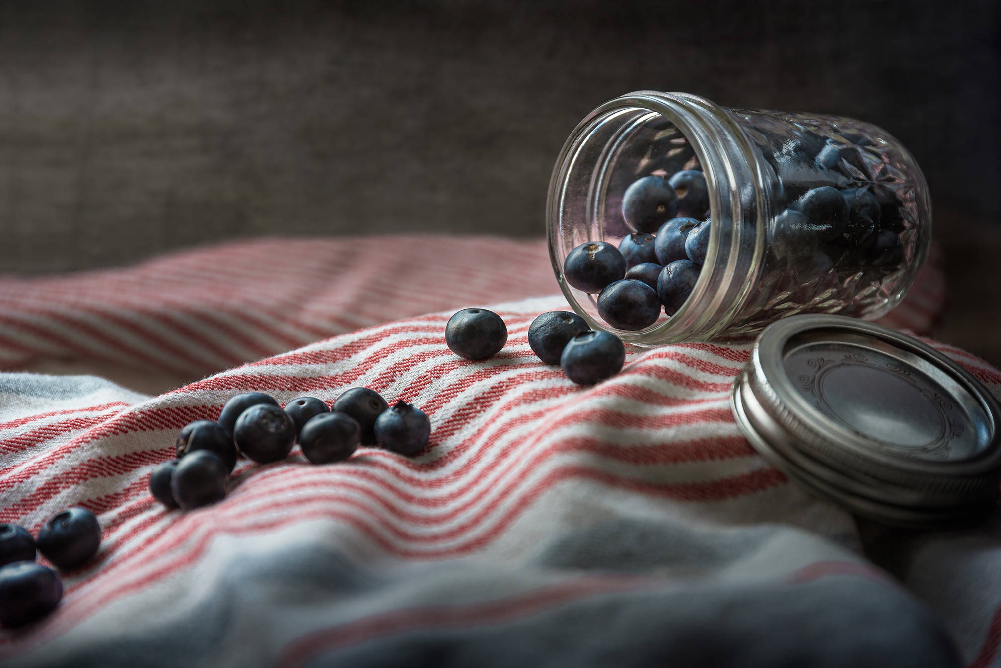 Blueberry 桌布 壁紙 — 新鮮藍莓從玻璃罐中溢出，落在紅白條紋布上，柔和自然光線下。
