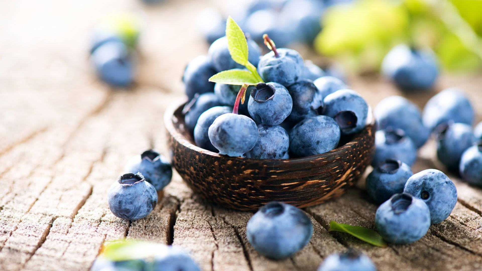 Fruits 桌布 壁紙 — 新鮮藍莓放在椰子殼碗中，置於經風化的木質表面上，周圍散落著莓果和綠葉。
