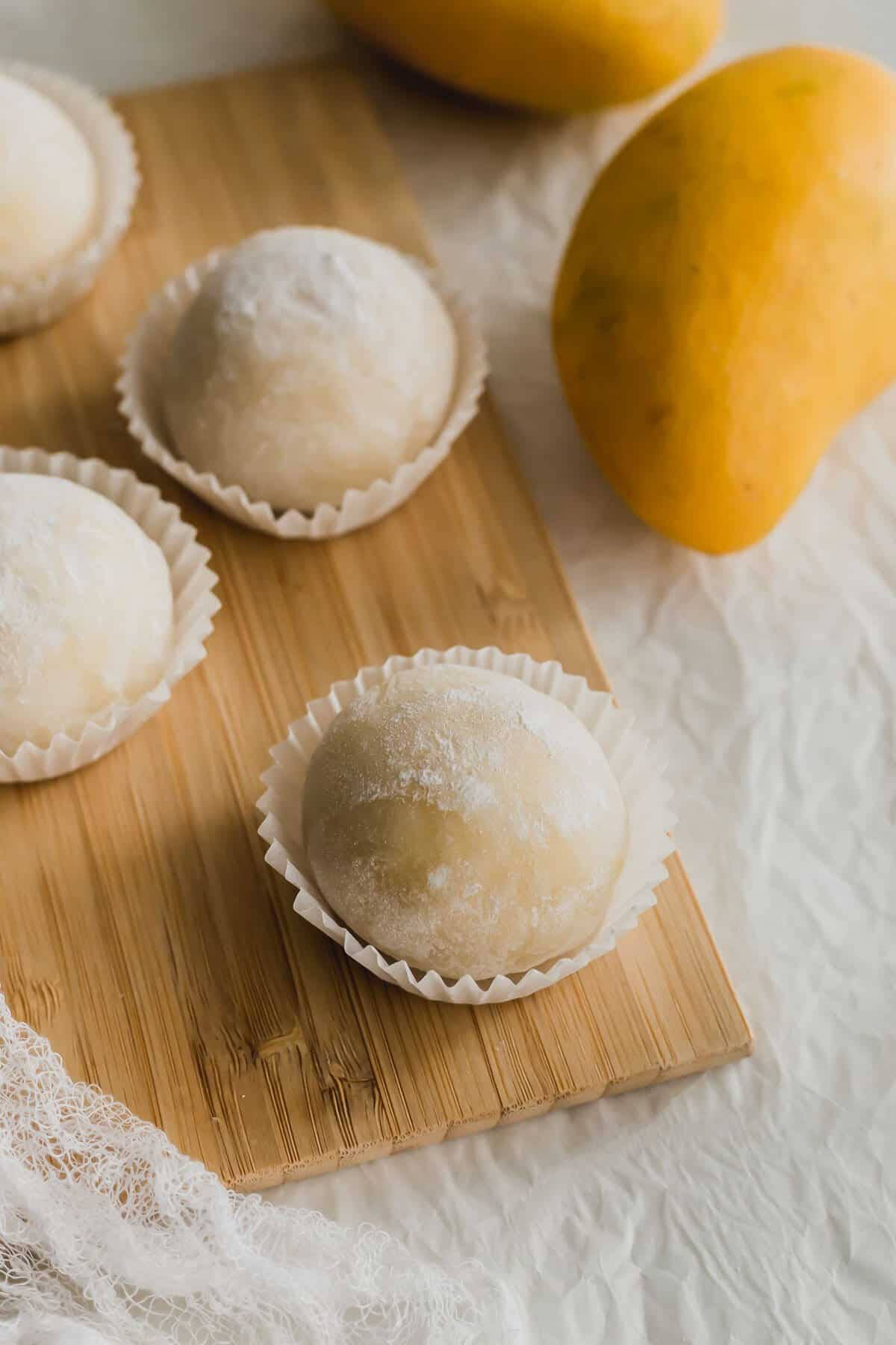 Mochi 桌布 壁紙 — 新鮮製作的芒果麻糬球撒上糖粉，放在紙杯中，置於木板上，旁邊有完整的芒果。