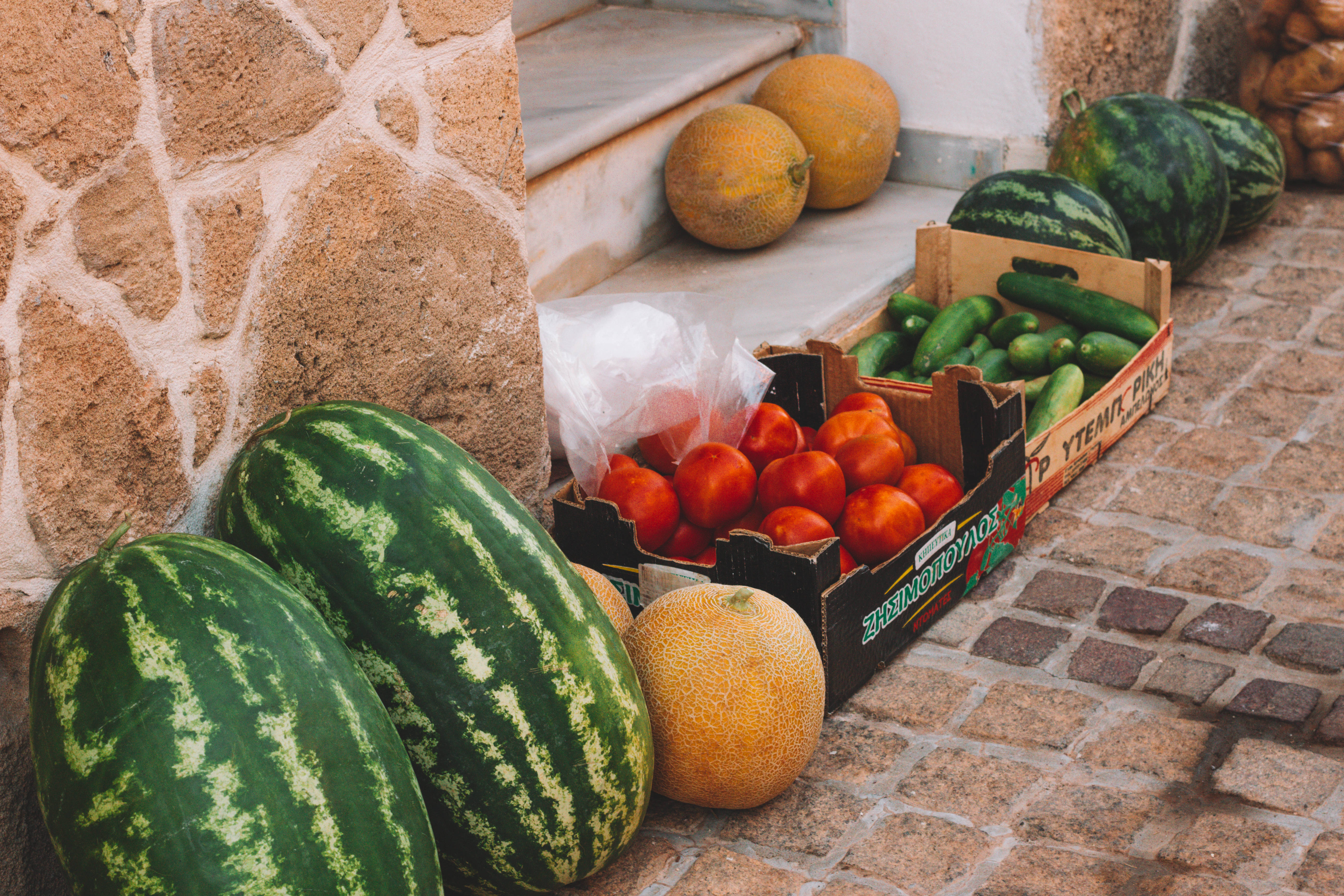 Watermelon 桌布 壁紙 — 新鮮西瓜、哈密瓜和番茄擺放在木箱中，展示在陽光照耀的鵝卵石街道上，旁邊是石牆。