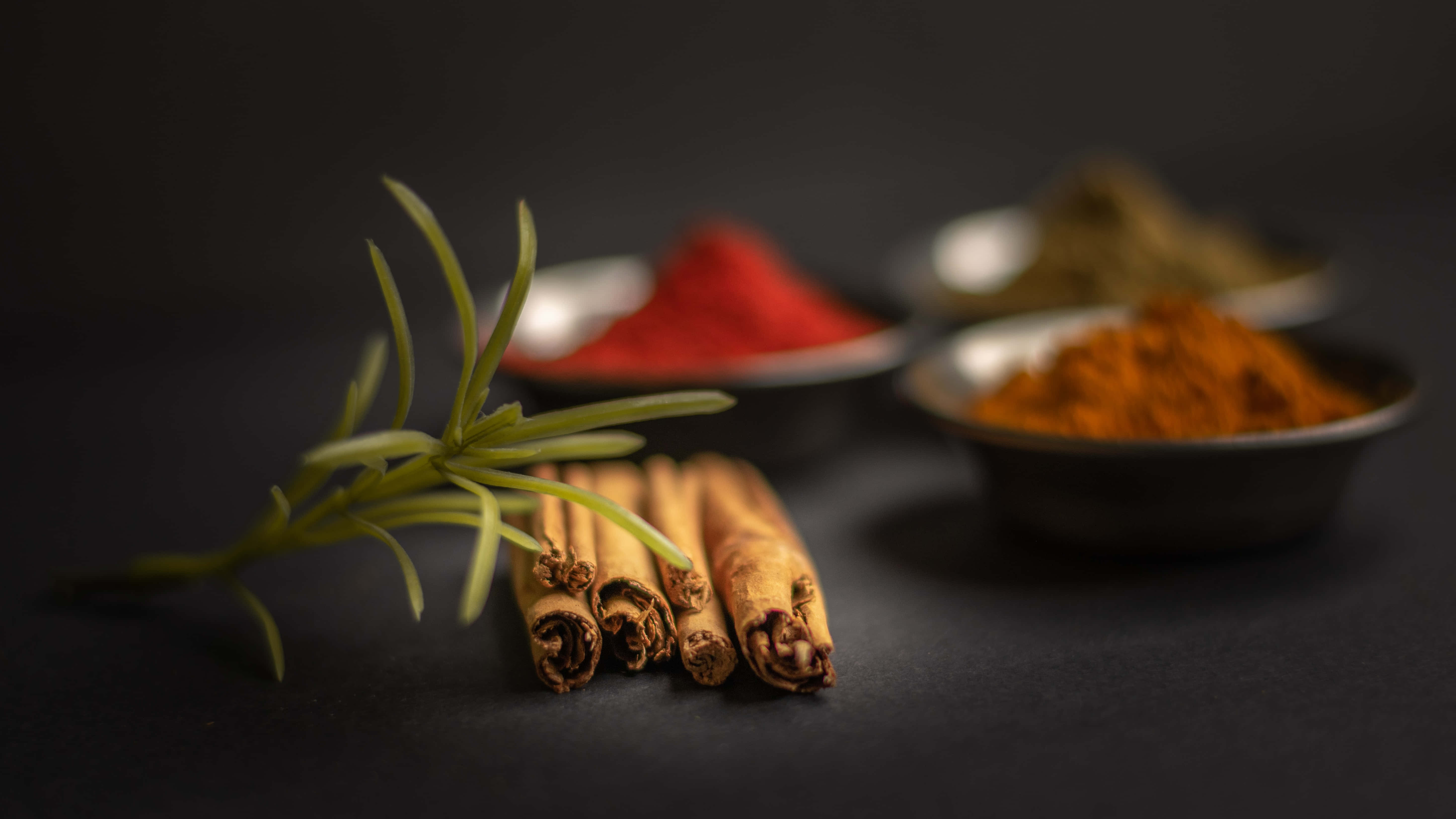 Spices 桌布 壁紙 — 新鮮迷迭香和肉桂棒放在深色表面上，旁邊是裝有鮮豔紅色和橙色香料的碗。