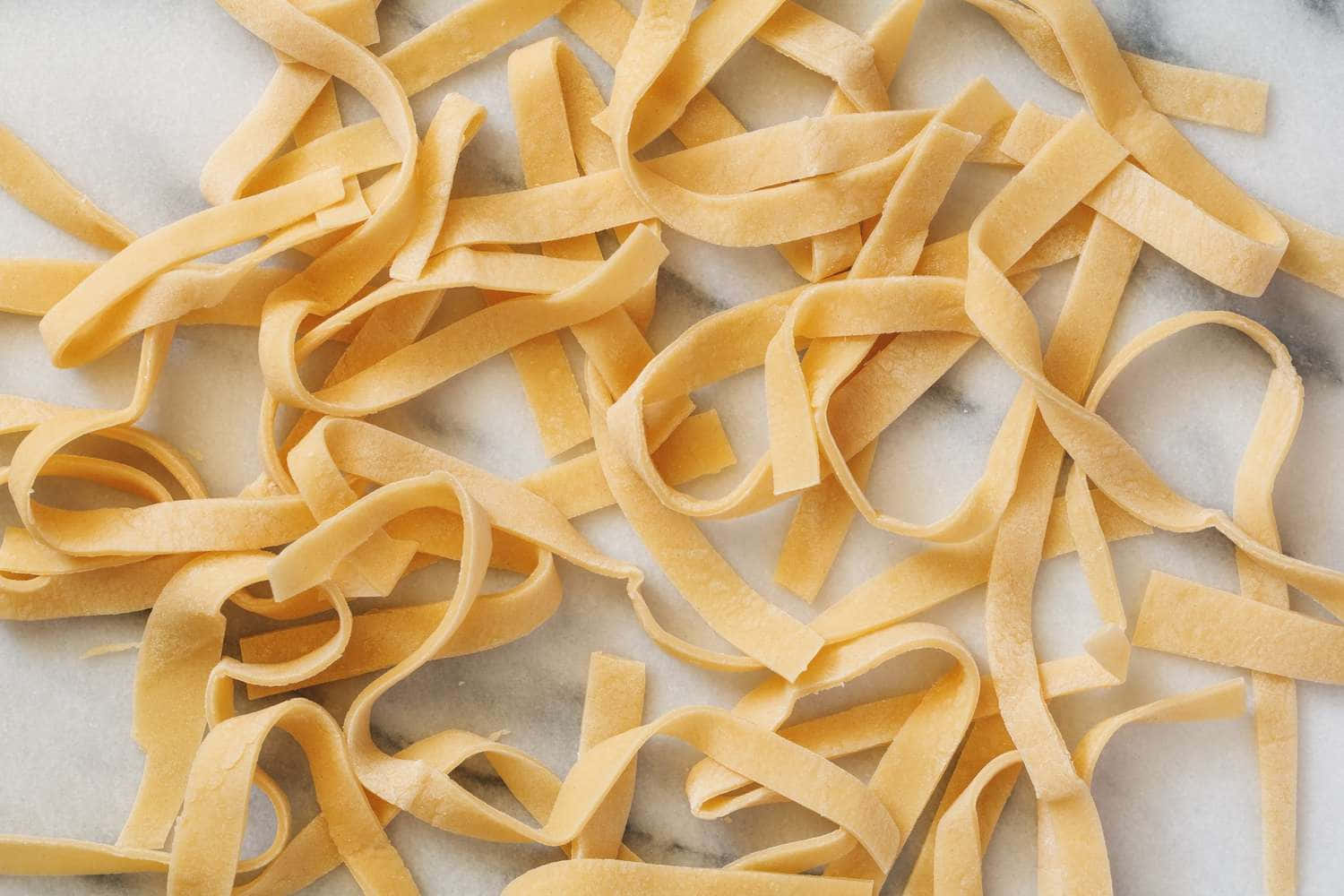 Noodles 桌布 壁紙 — 新鮮金黃色義大利麵絲帶散落在清涼的大理石表面上，柔和自然光線下。