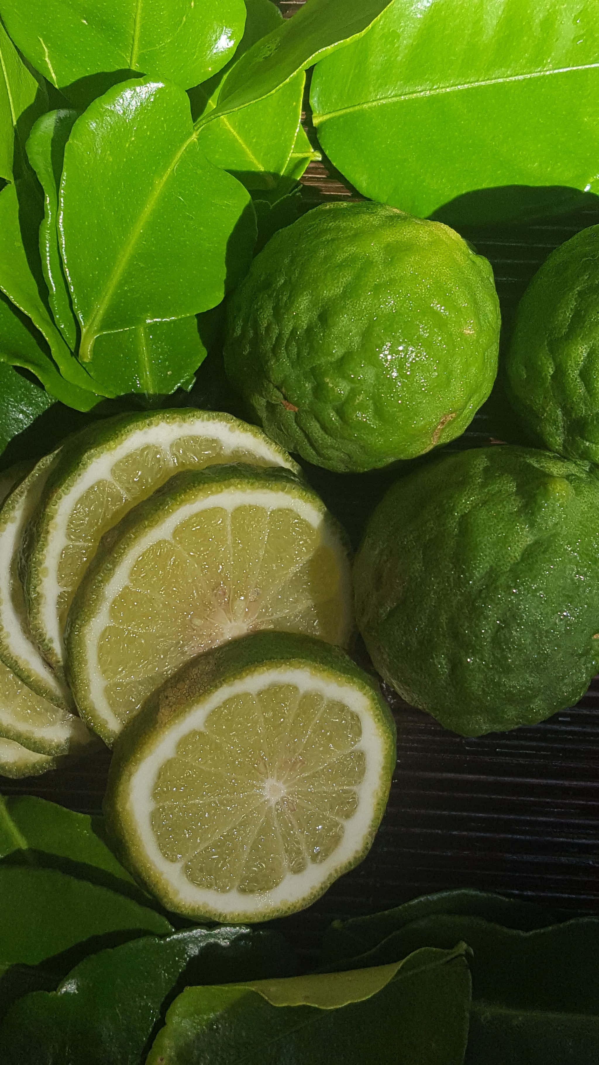 Fruit 桌布 壁紙 — 新鮮青檸切片和完整青檸，依偎在光澤的熱帶葉子中，放在深色木質表面上。