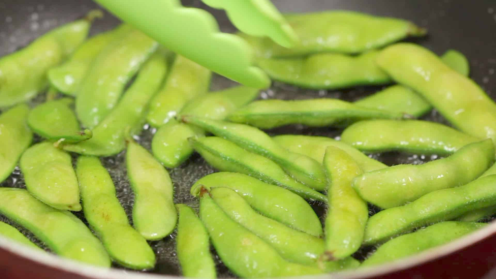 beans 桌布 壁紙 — 新鮮青毛豆莢在熱油中滋滋作響，綠色夾子從紅色煎鍋中夾起一個豆莢。