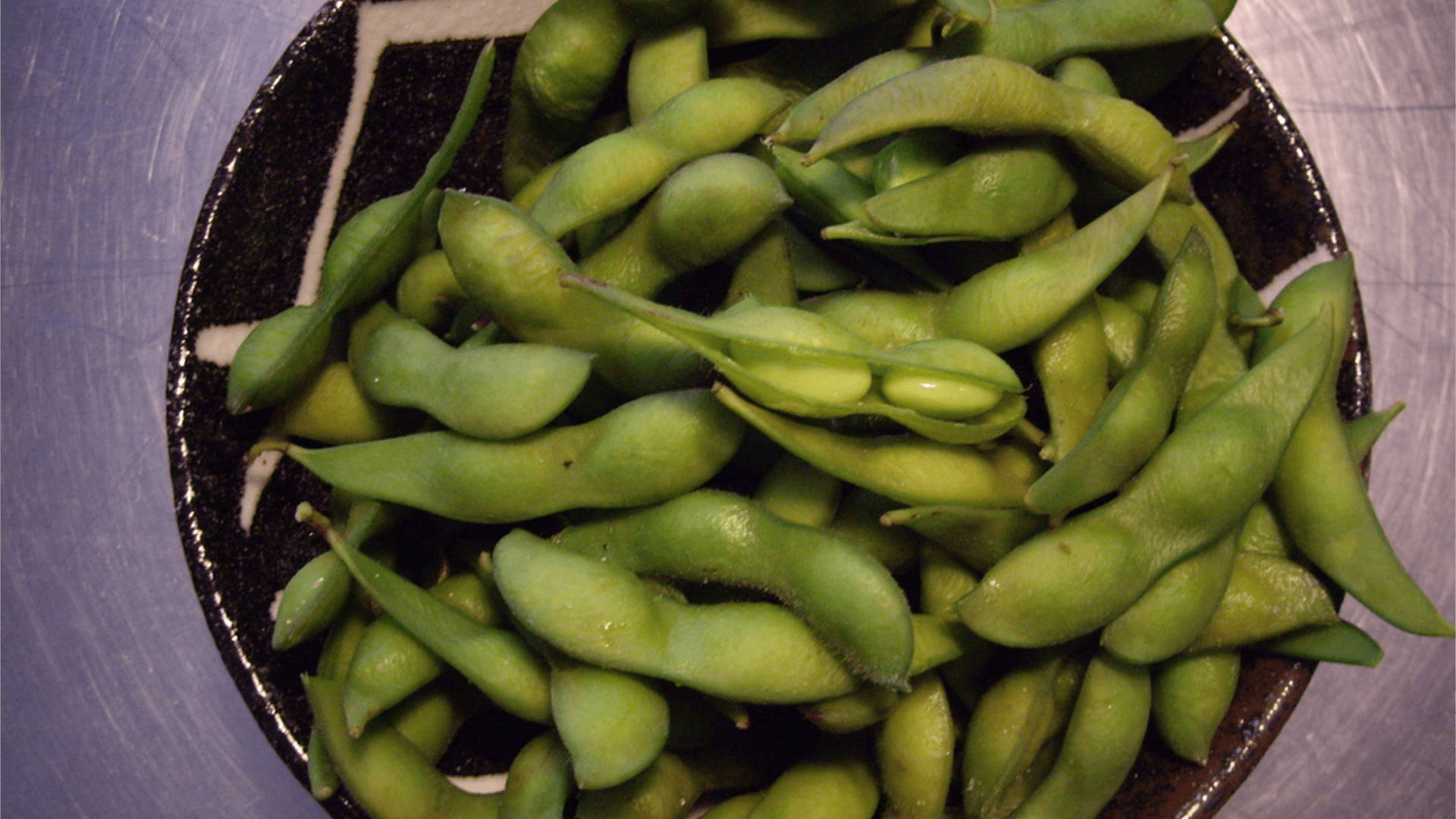 beans 桌布 壁紙 — 新鮮青毛豆莢堆疊在黑色斑點碗中，置於拉絲金屬表面上的特寫。