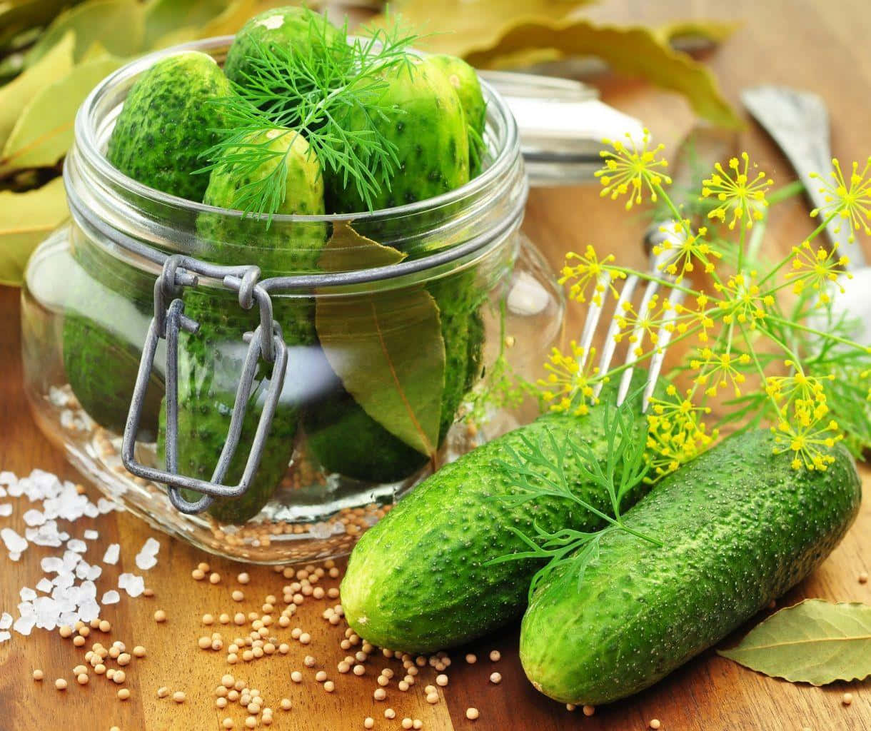 cucumber 桌布 壁紙 — 新鮮青瓜酸在玻璃罐中，配有蒔蘿和月桂葉，周圍是芥末籽、鹽和黃色花朵，置於木質表面上。