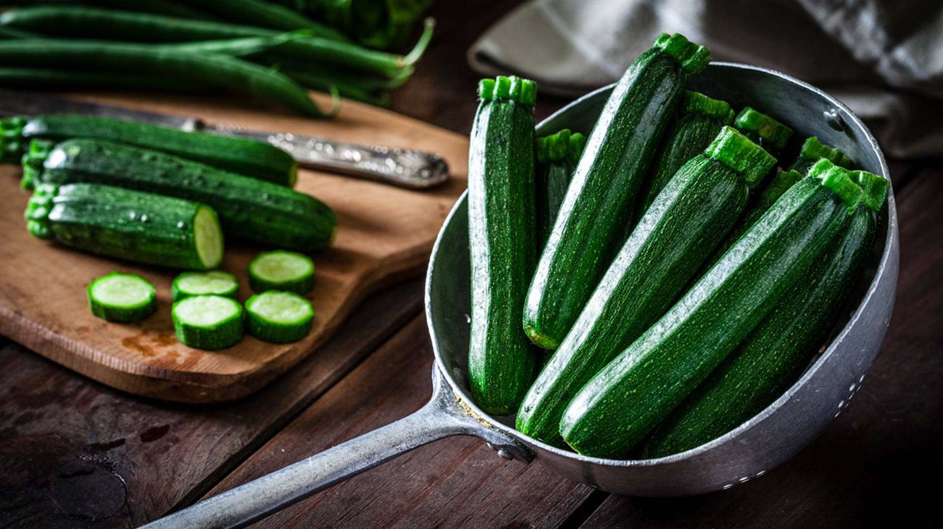 Food Photography 桌布 壁紙 — 新鮮青綠的櫛瓜堆在金屬濾盆中，旁邊木砧板上放有切片。