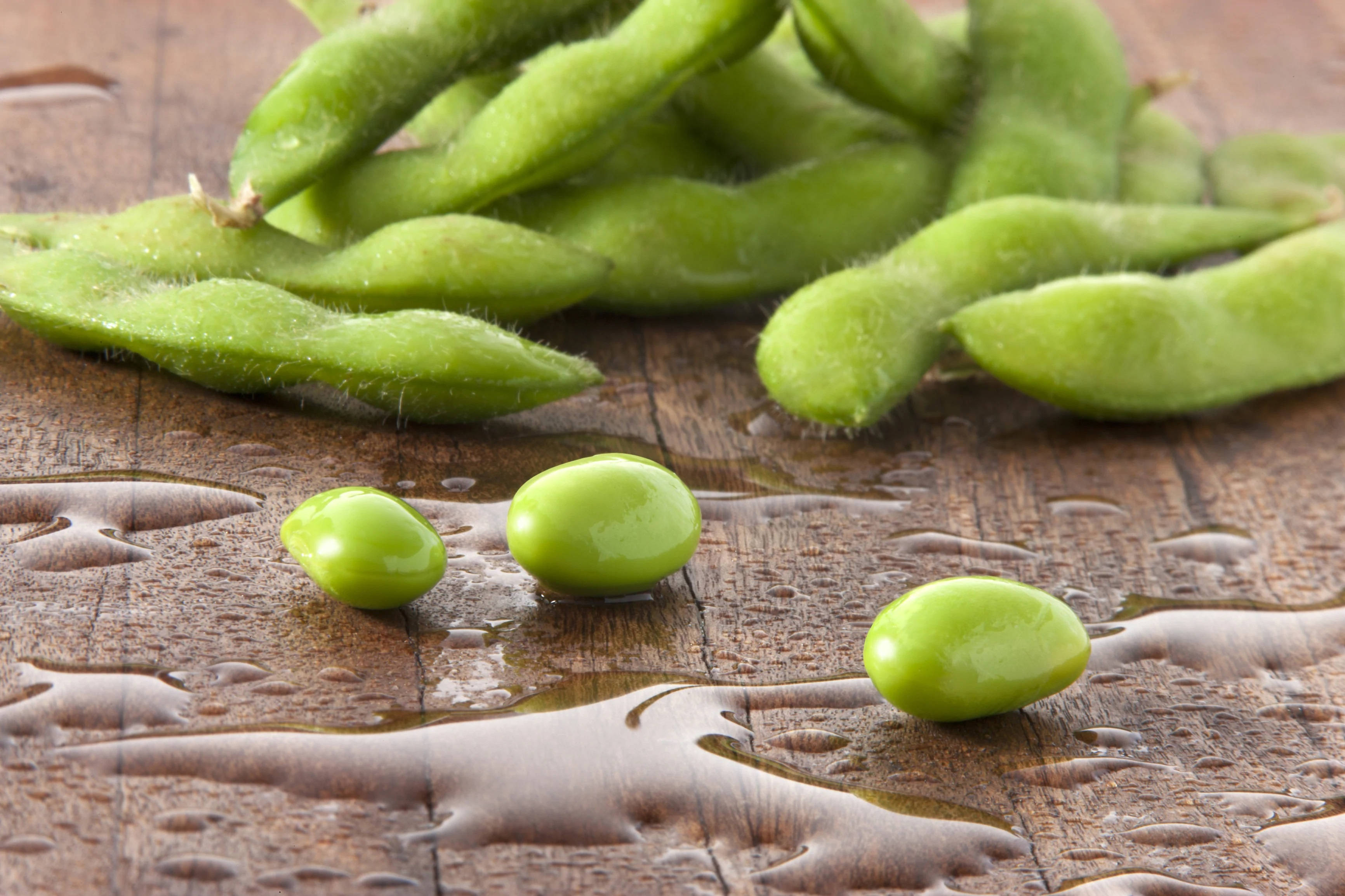 Vegetables 桌布 壁紙 — 新鮮青綠的毛豆從豆莢中灑落在潮濕的木質表面上，帶有閃亮的水珠。