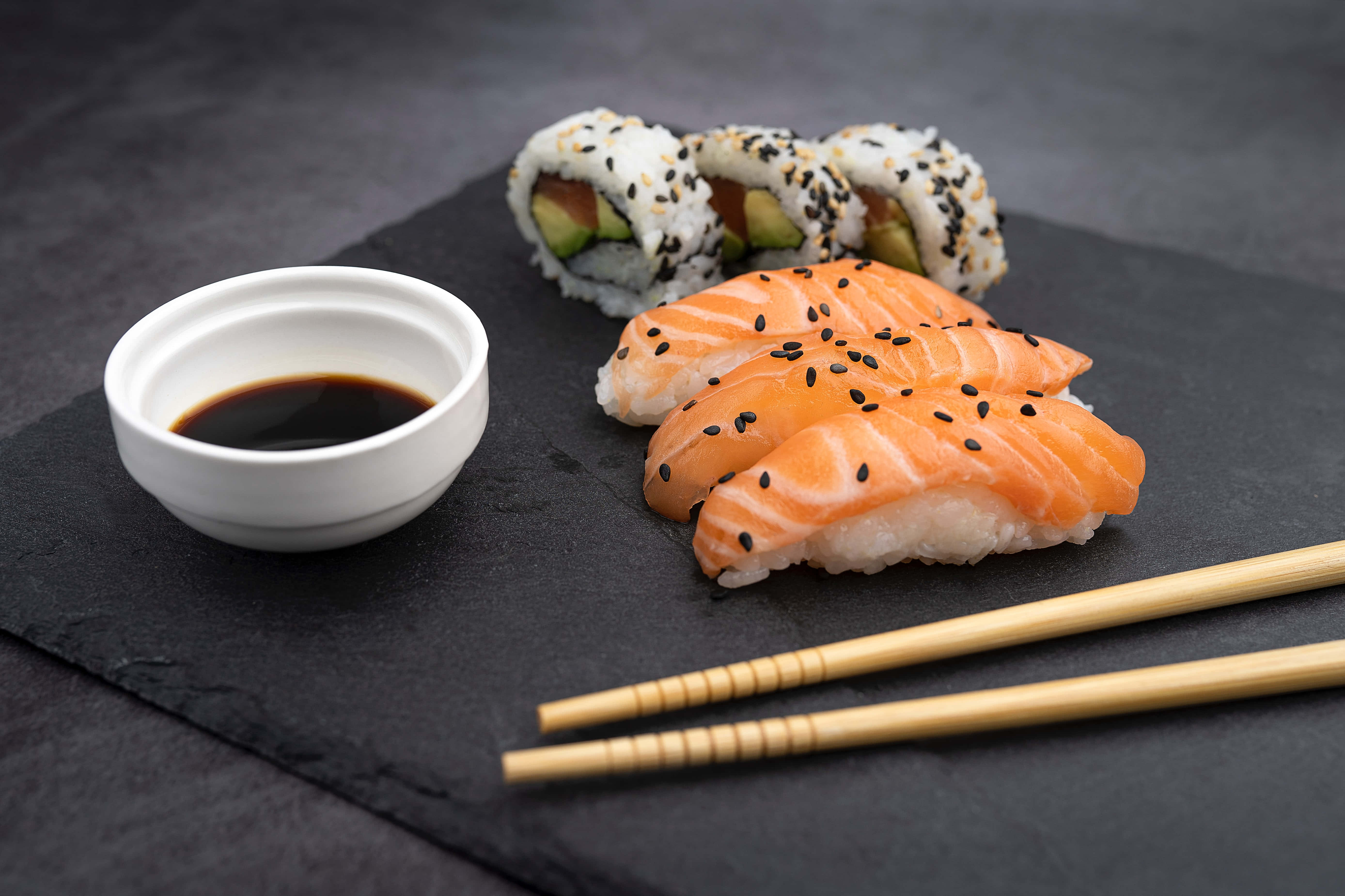Sushi 桌布 壁紙 — 新鮮鮭魚握壽司和酪梨卷，飾以黑芝麻，旁邊有醬油和筷子，放在深色石板盤上。