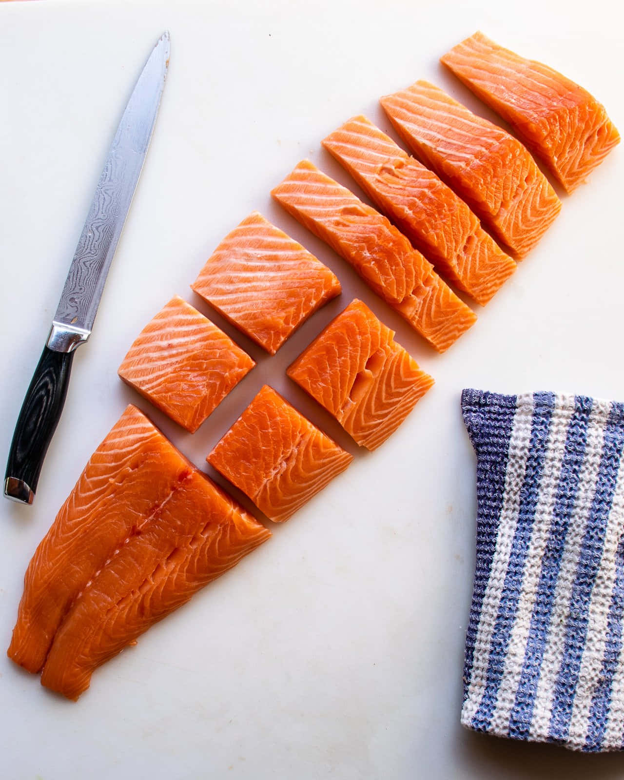 Salmon 桌布 壁紙 — 新鮮鮭魚柳切成塊，放在白色砧板上，旁邊有刀和條紋布。