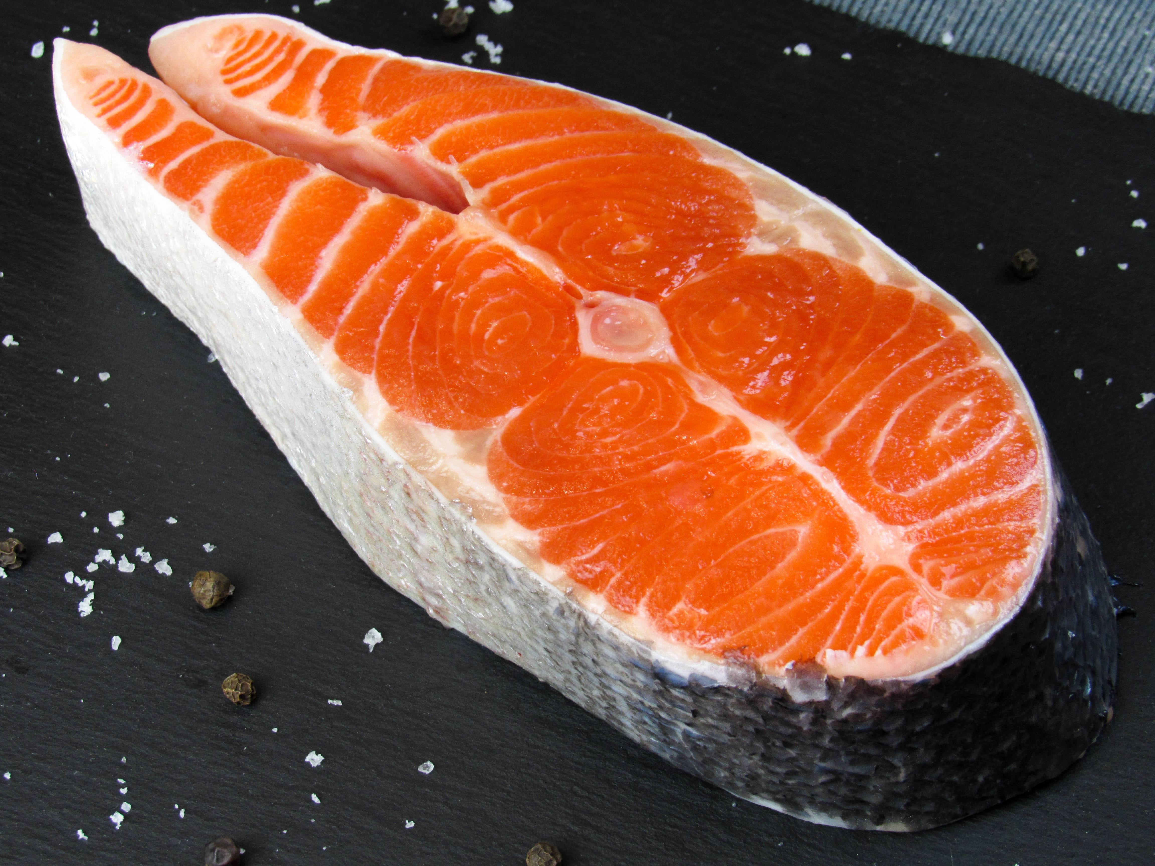 meat 桌布 壁紙 — 新鮮鮭魚片，帶有鮮豔橙色肉質和白色大理石紋，放在灑有粗鹽和胡椒粒的深色石板表面上。