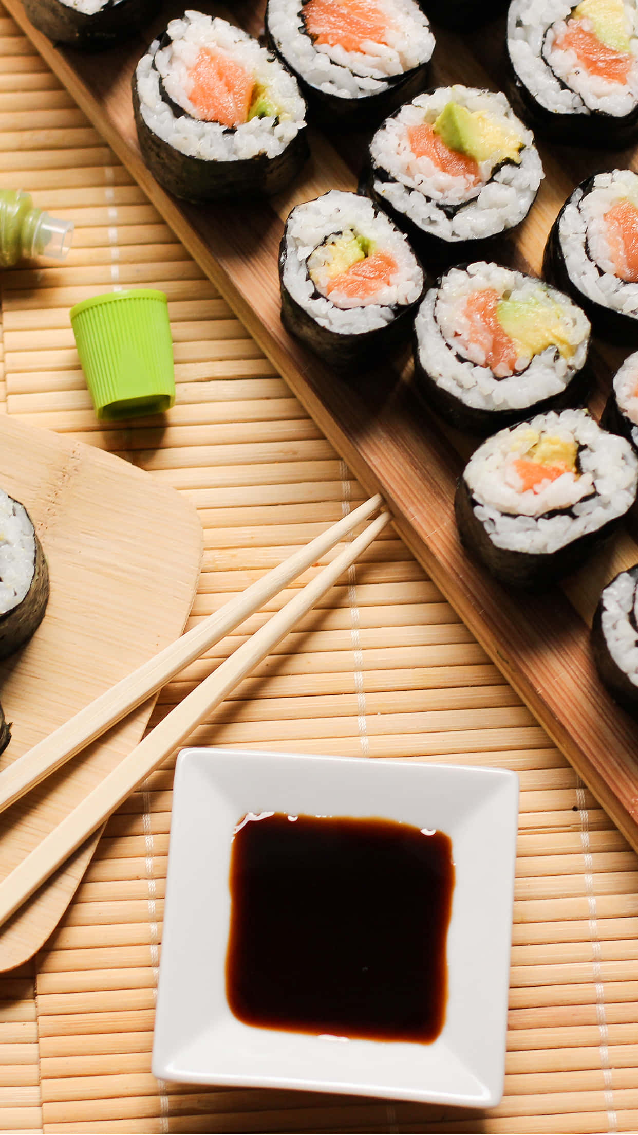 Sushi 桌布 壁紙 — 新鮮鮭魚牛油果壽司卷擺放在木托盤上，旁邊有醬油和筷子在竹蓆上。