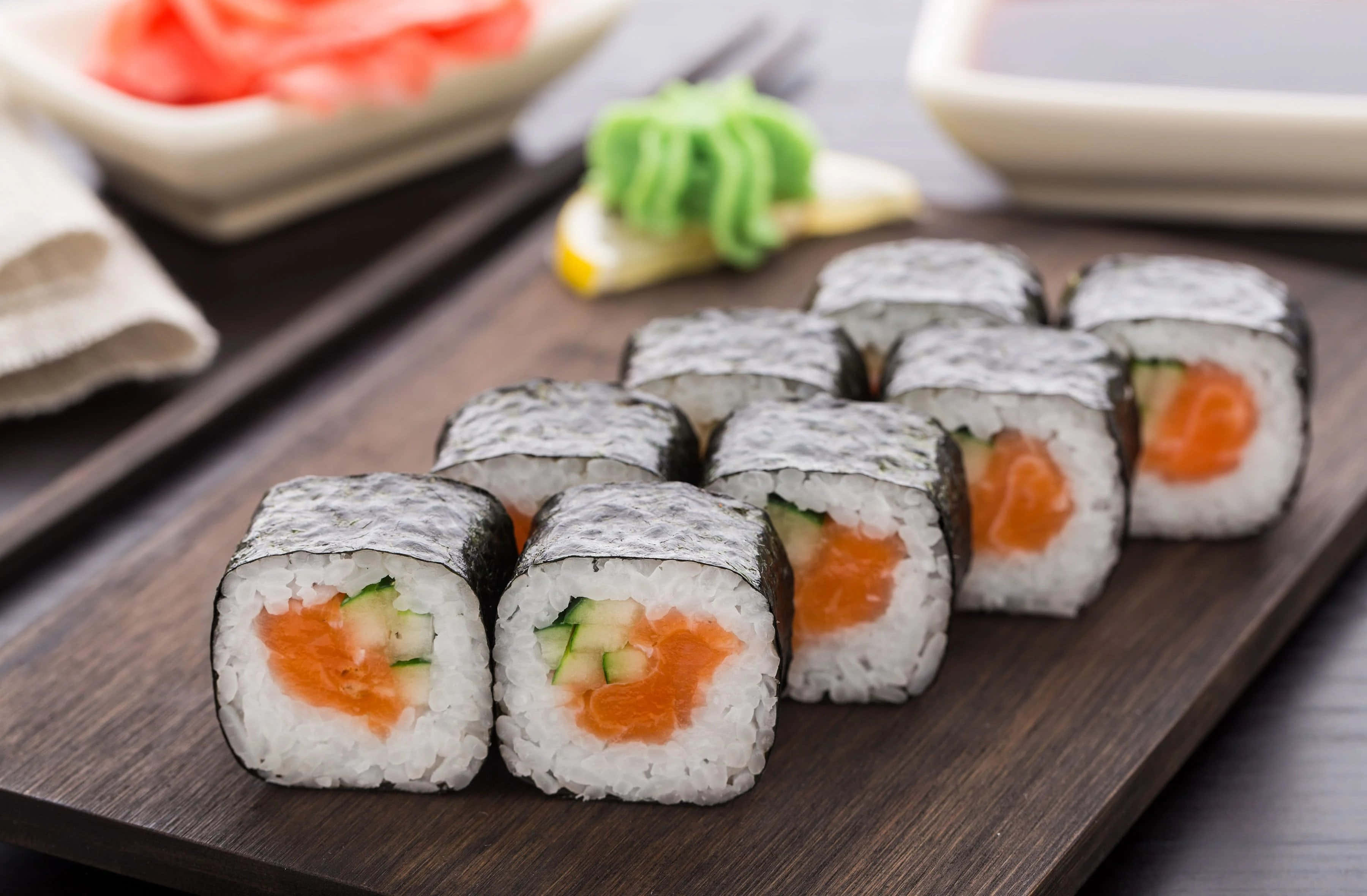 Sushi 桌布 壁紙 — 新鮮鮭魚黃瓜壽司卷整齊排列在深色木製托盤上，背景有芥末和薑。