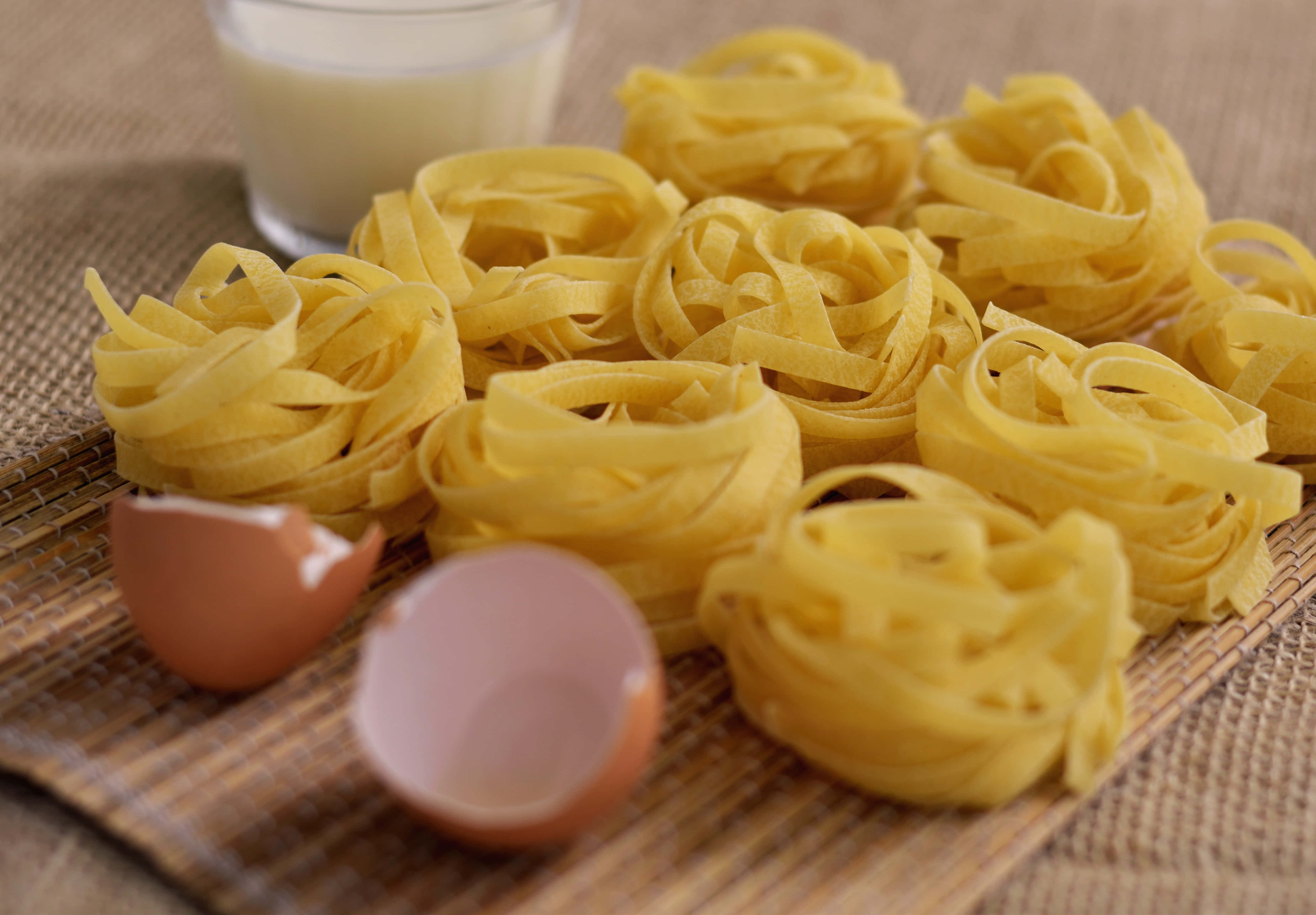 Brown Eggs 桌布 壁紙 — 新鮮黃色義大利麵巢排列在編織墊上，旁邊有破碎的蛋殼和一杯牛奶。