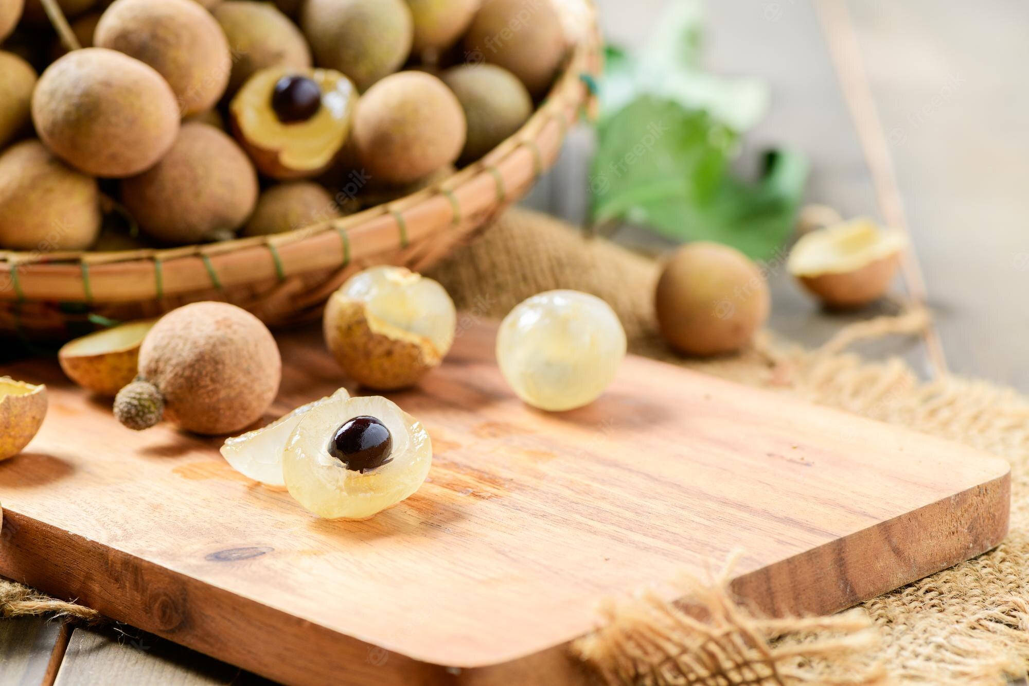 Fruits 桌布 壁紙 — 新鮮龍眼果實，帶有半透明白色果肉和深色種子，放在木製砧板上，旁邊有一個編織籃子。