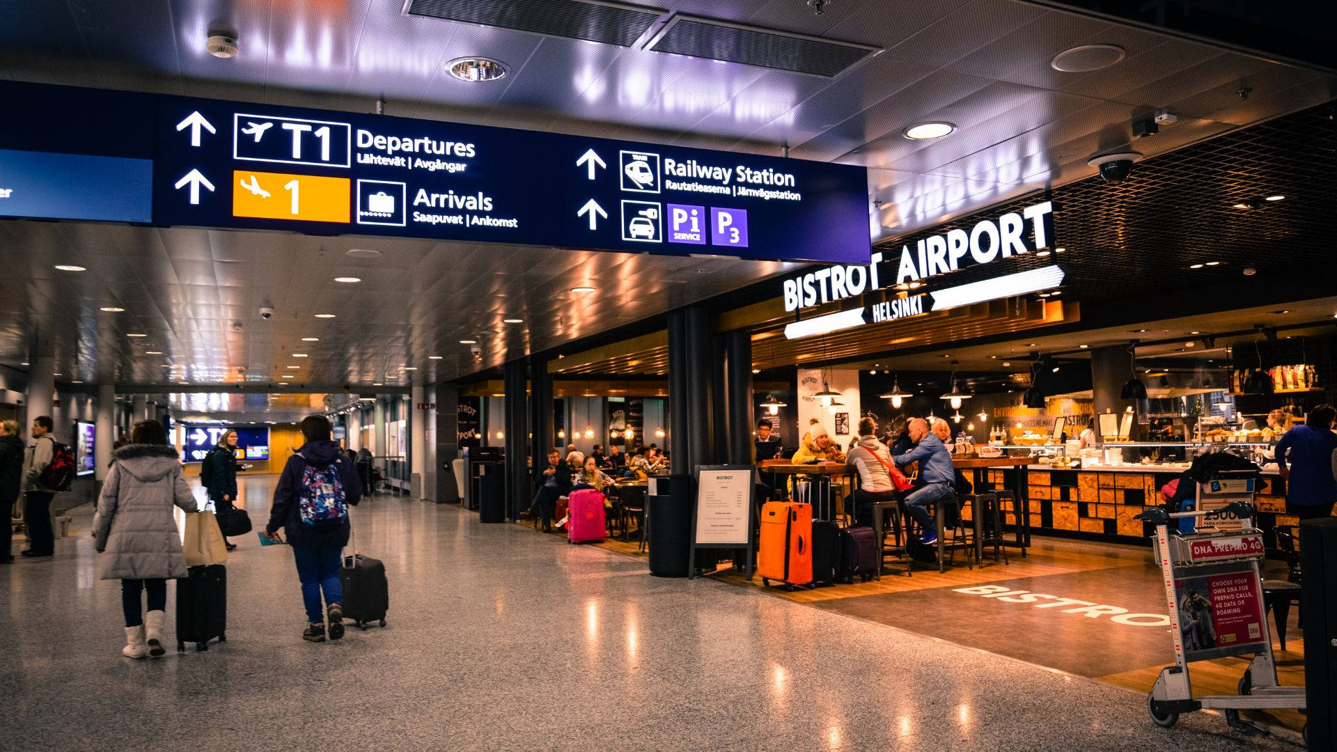 Airport 桌布 壁紙 — 旅客在明亮的標誌和熱鬧的小餐館下，於溫暖的人造燈光中穿梭於繁忙的機場航廈。
