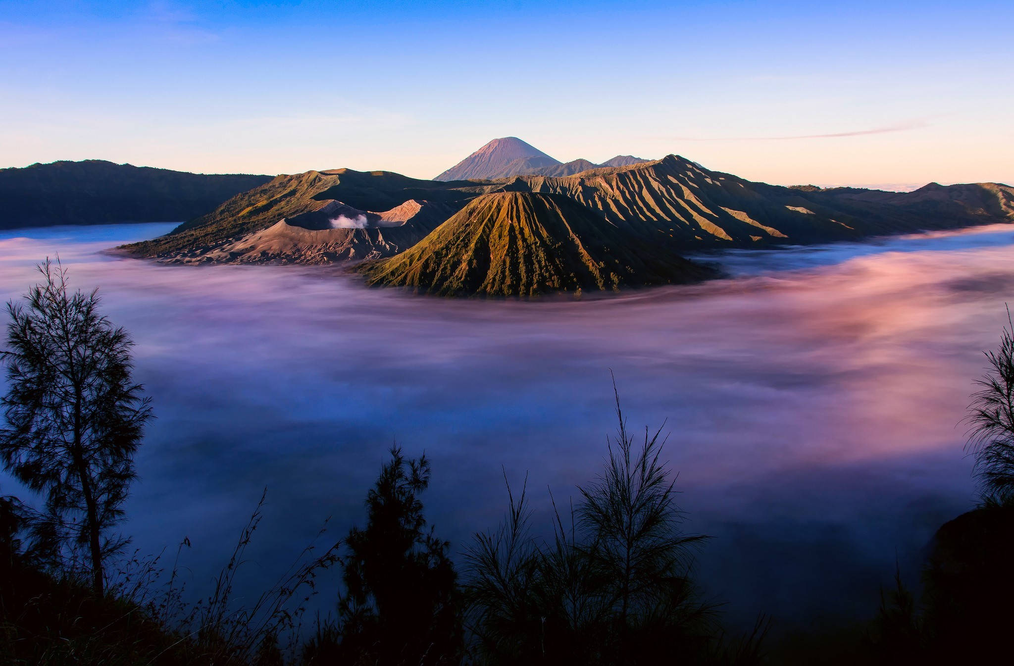 印尼 桌布 壁紙 — 日出以金色光芒沐浴火山峰頂，霧雲在下方旋轉，宛如紫色和藍色的海洋。
