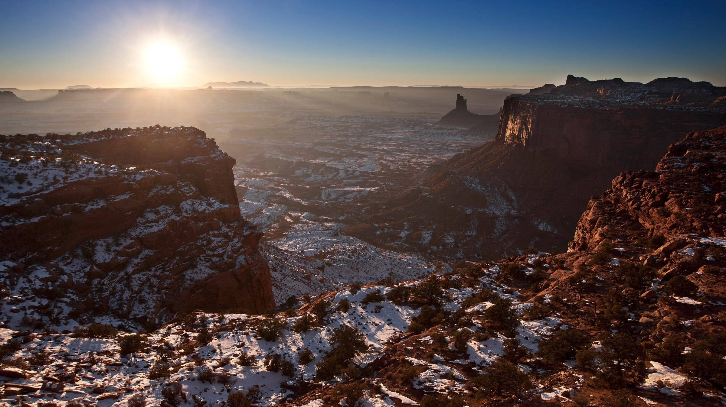 Canyonlands National Park 桌布 壁紙 — 日出在白雪覆蓋的峽谷上灑下金色光芒，紅色岩層和遠處的平頂山在清澈的藍天下展現。