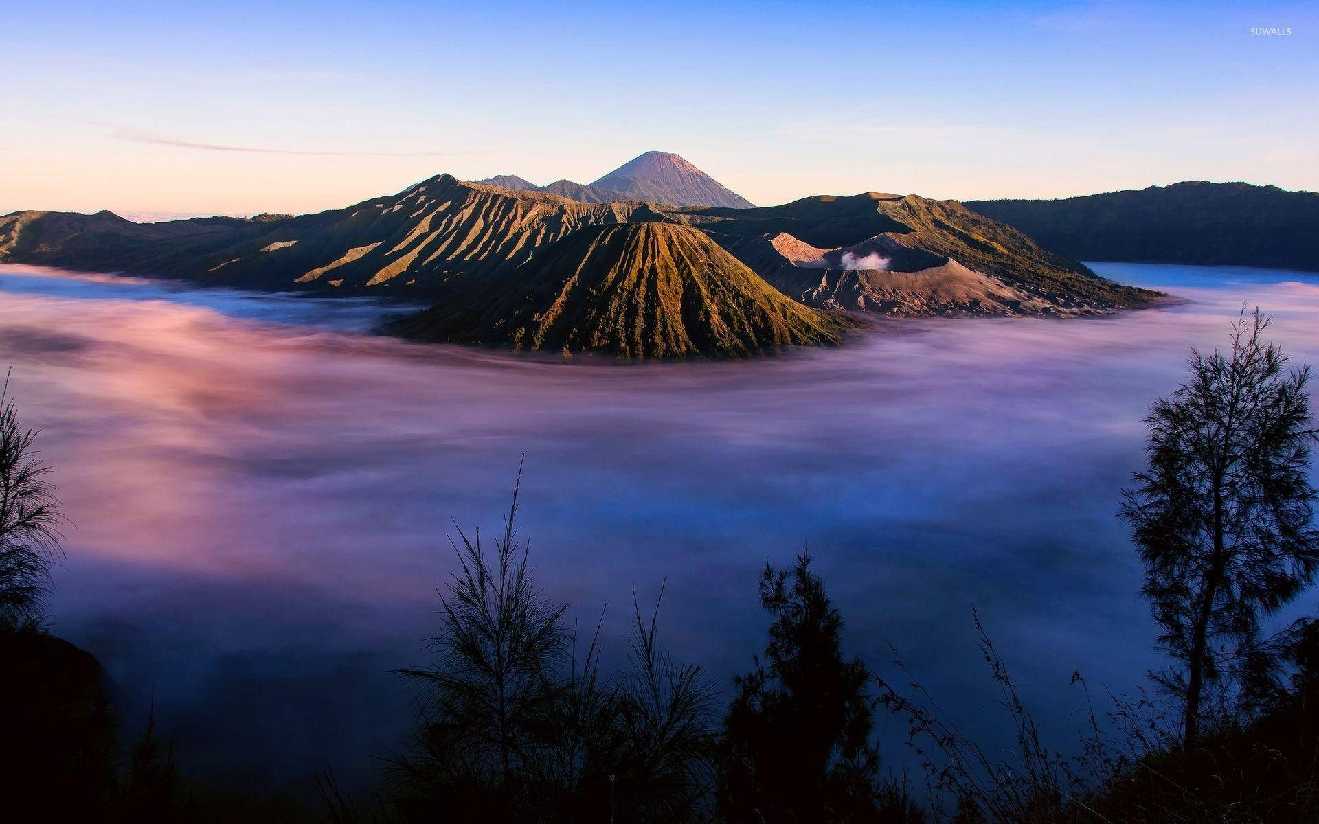 Jakarta 桌布 壁紙 — 日出將火山山峰沐浴在金色光芒中，霧氣雲朵在下方旋轉，像一片紫色和藍色的海洋。