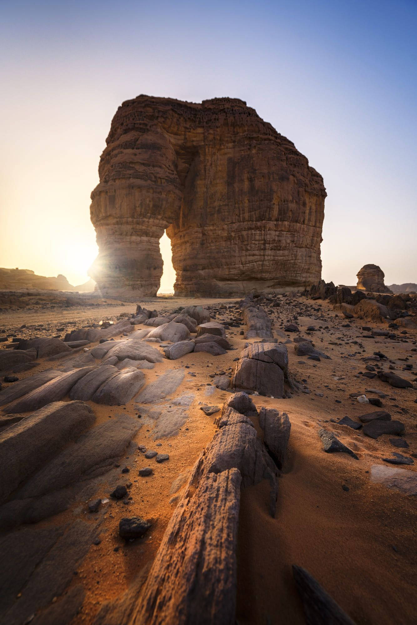 Saudi 桌布 壁紙 — 日出時，陽光穿過沙漠景觀中一個巨大的天然岩石拱門，在岩石地形上投下長長的陰影。