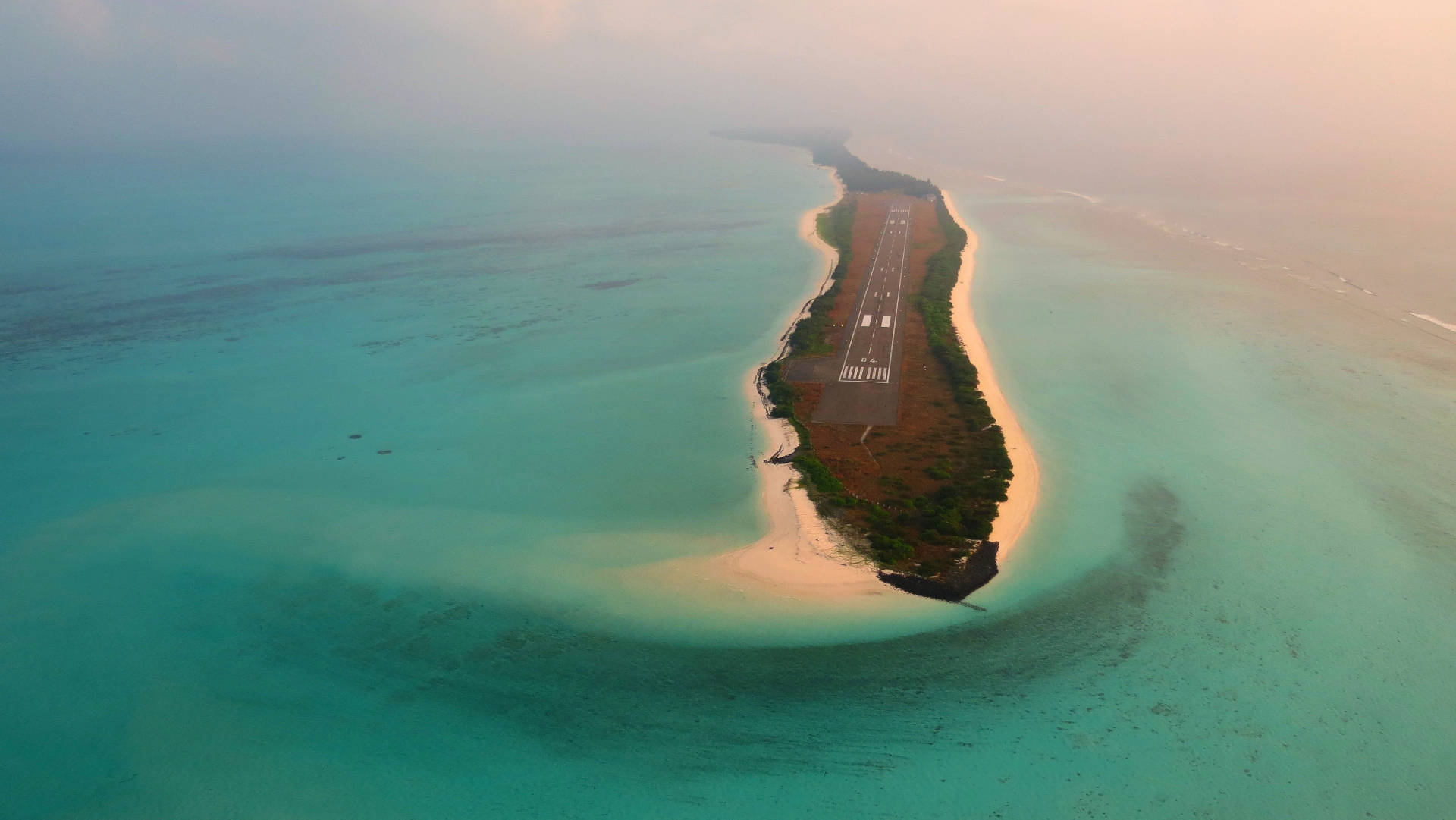 Runway 桌布 壁紙 — 日出時分，一條跑道穿過茂密熱帶島嶼的鳥瞰圖，周圍環繞著碧綠海水。