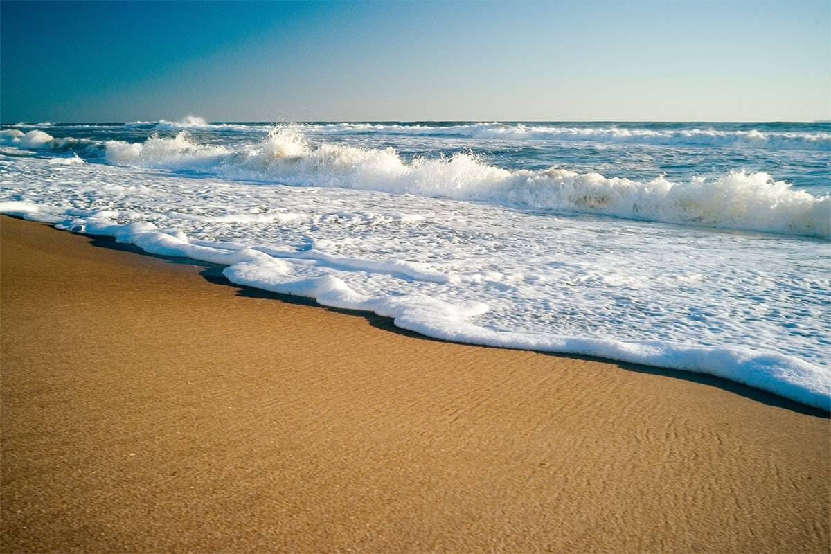 海灘 桌布 壁紙 — 日出時分，平靜的海浪在清澈的藍天下拍打著金色的沙灘。
