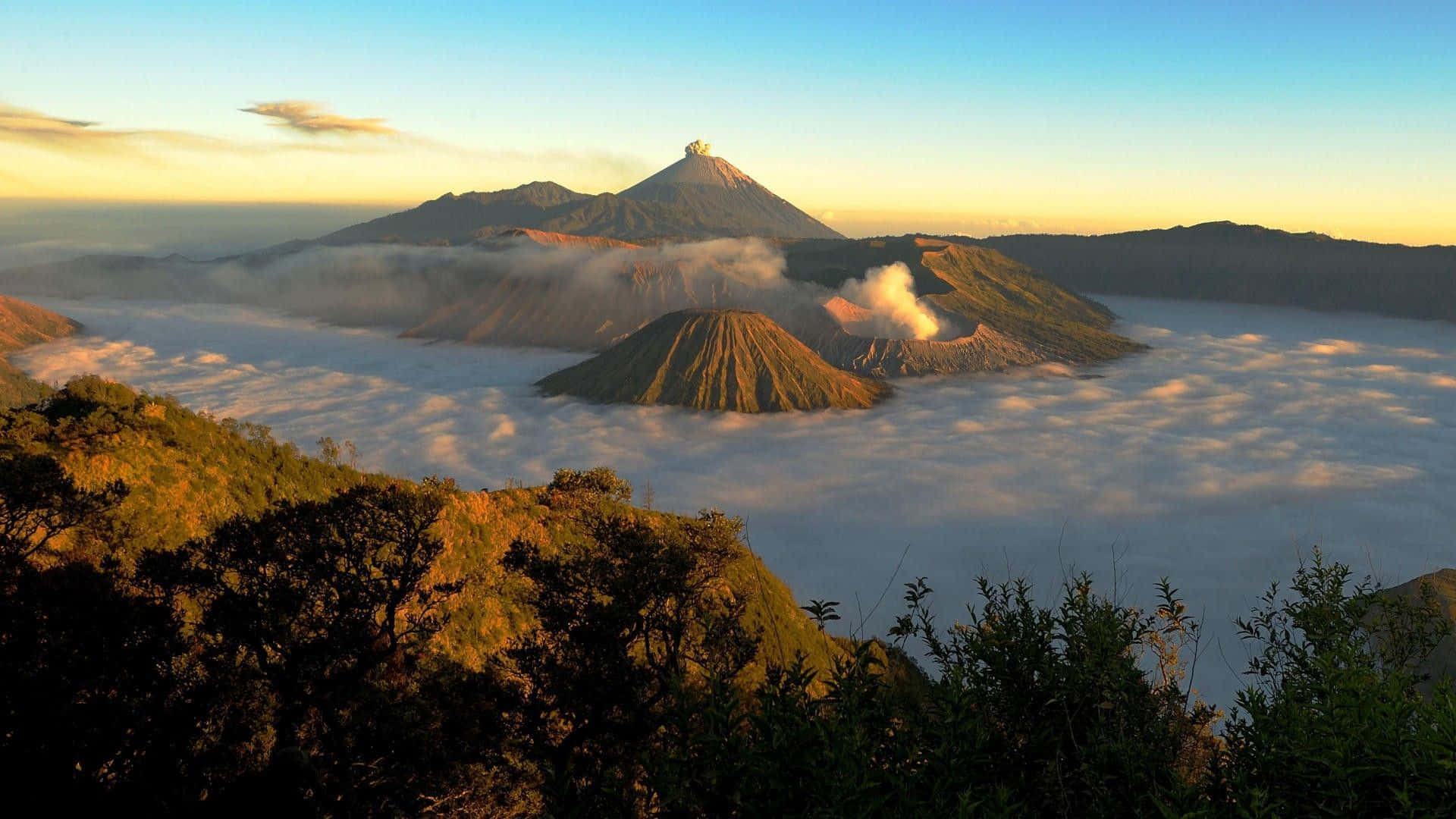 印尼 桌布 壁紙 — 日出時分，雄偉的火山山脈聳立在雲海之上，其中一座火山正活躍地噴出煙霧。