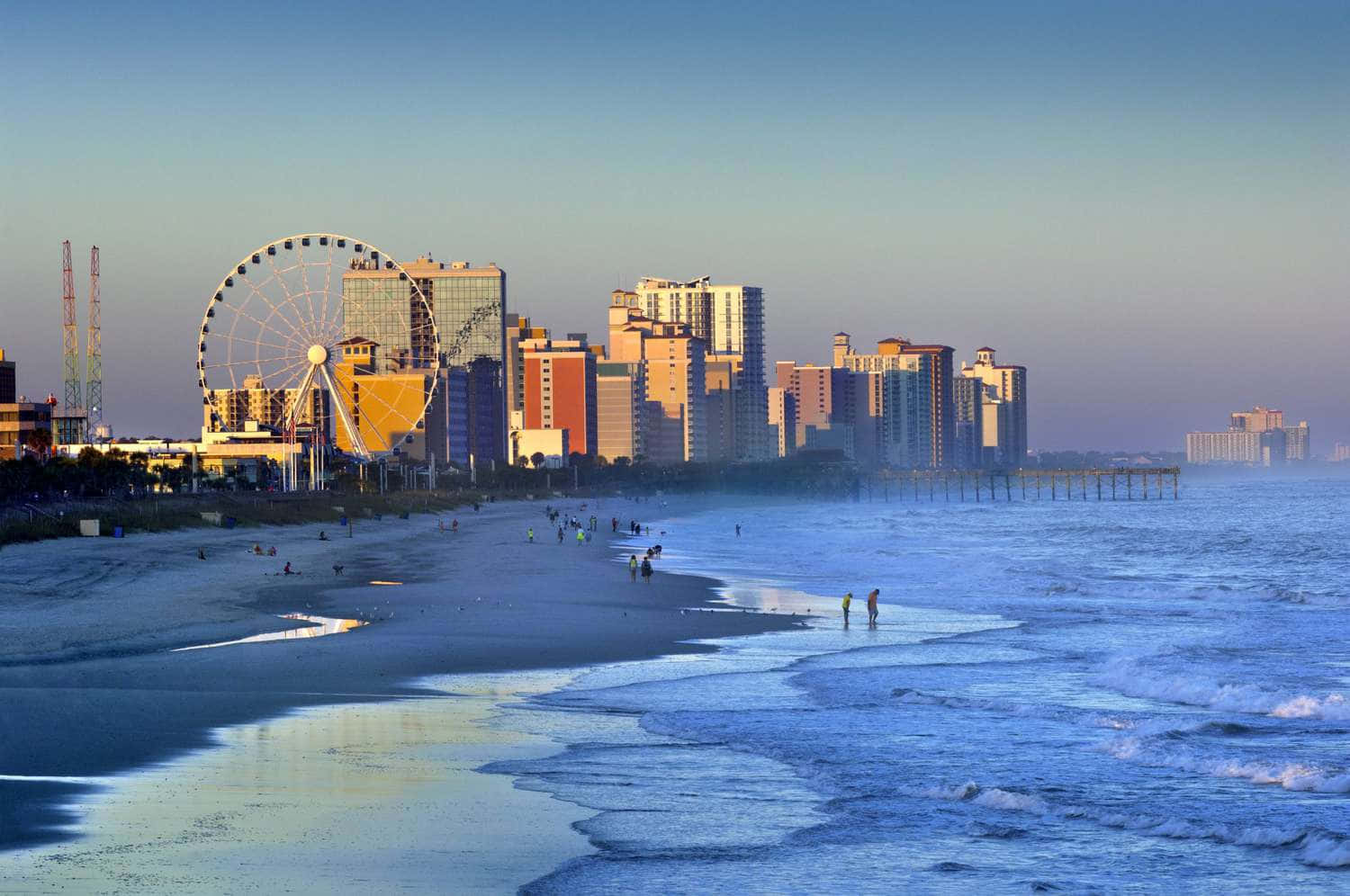 Myrtle Beach 桌布 壁紙 — 日出時分的沿海城市天際線，一座巨大的摩天輪聳立在海灘遊客和拍打的海浪之上。