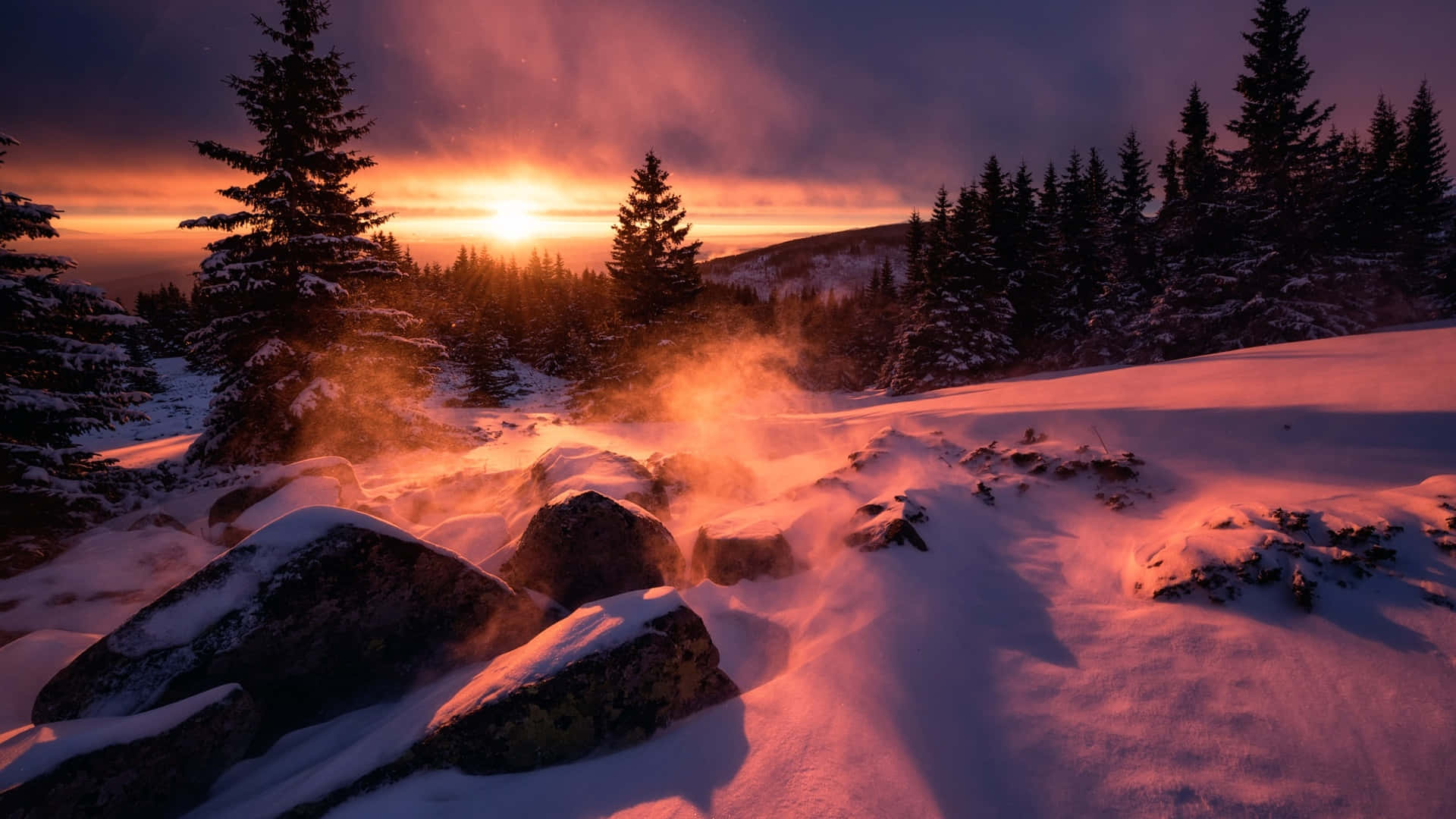 1920x1080 Winter 桌布 壁紙 — 日出時分的雪覆蓋山景，岩石上升起蒸氣，將溫暖的橙色光線投射在積雪的常青樹上。
