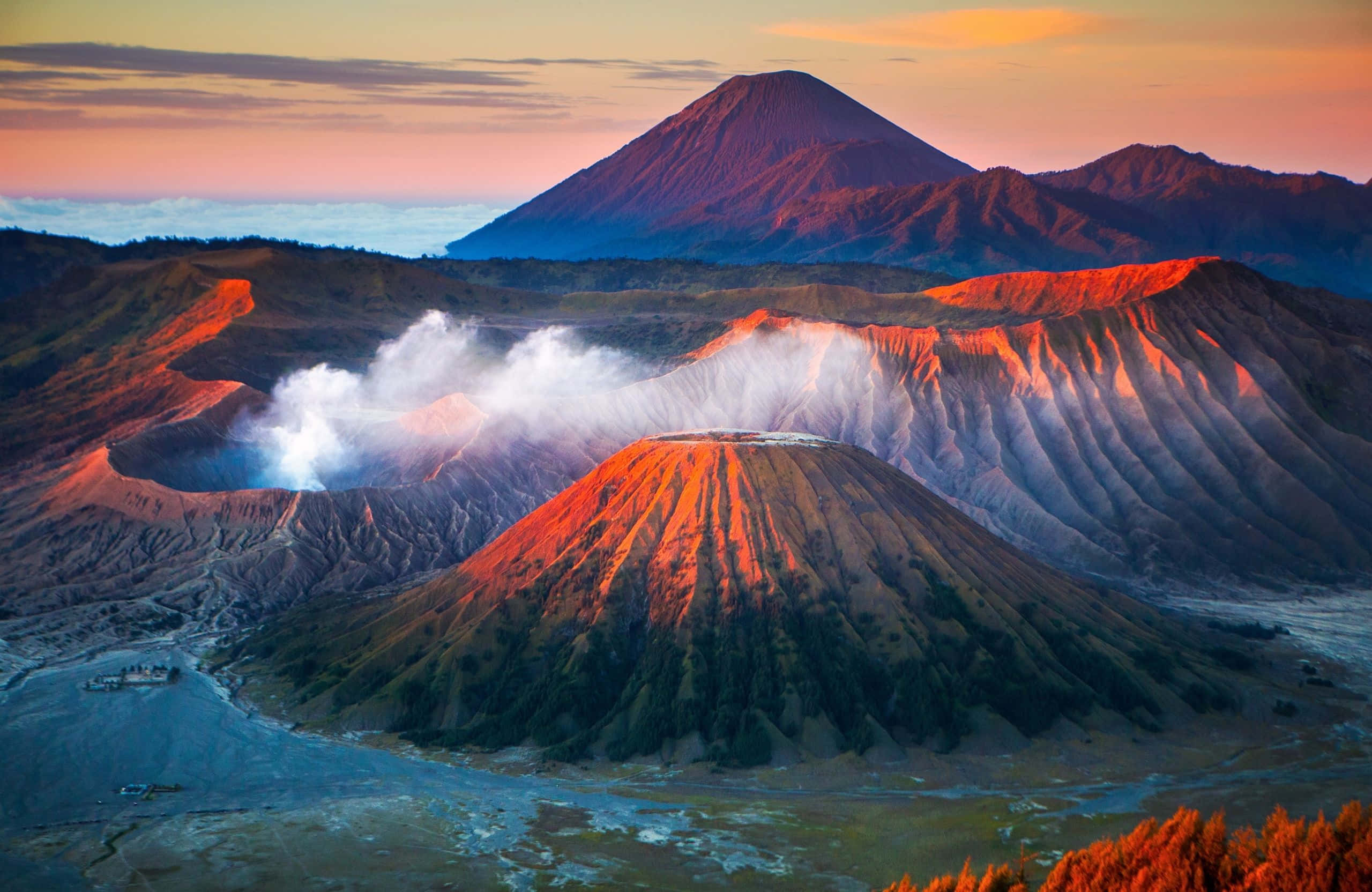 2560x1664 桌布 壁紙 — 日出時的火山景觀，蒸氣從火山口升起，山脈沐浴在溫暖的橙色和粉紅色光線中。