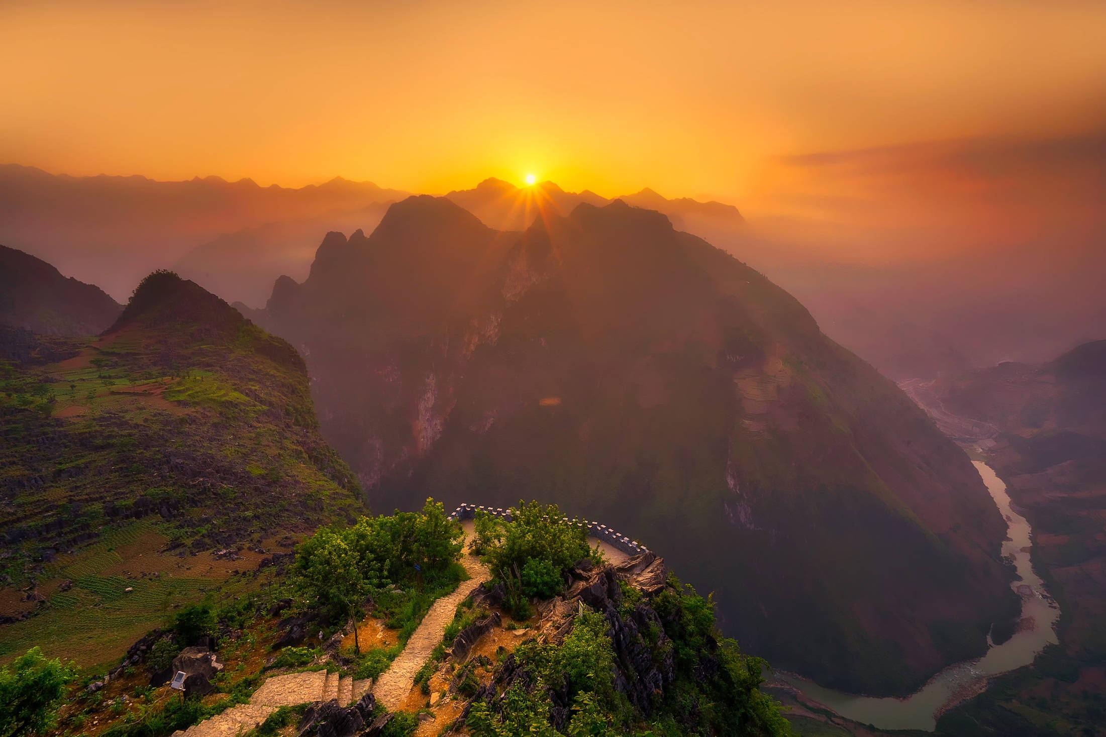 Vietnam 桌布 壁紙 — 日出籠罩著霧氣的山峰，下方有蜿蜒的河流和一條通往山谷的石徑。