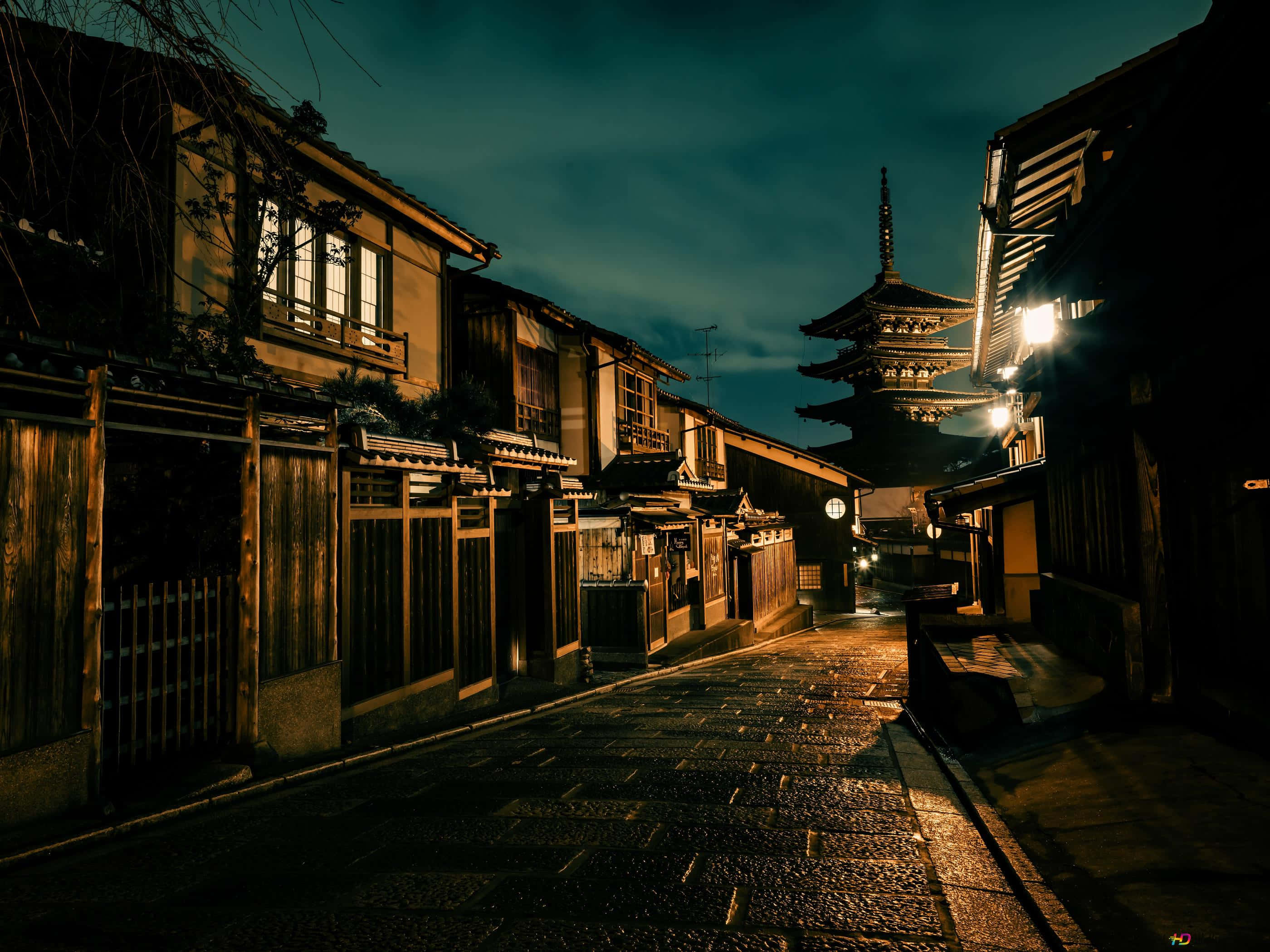 Night Street 桌布 壁紙 — 日本京都夜晚一條狹窄濕滑的石巷，被溫暖的燈籠照亮，遠處可見一座塔。