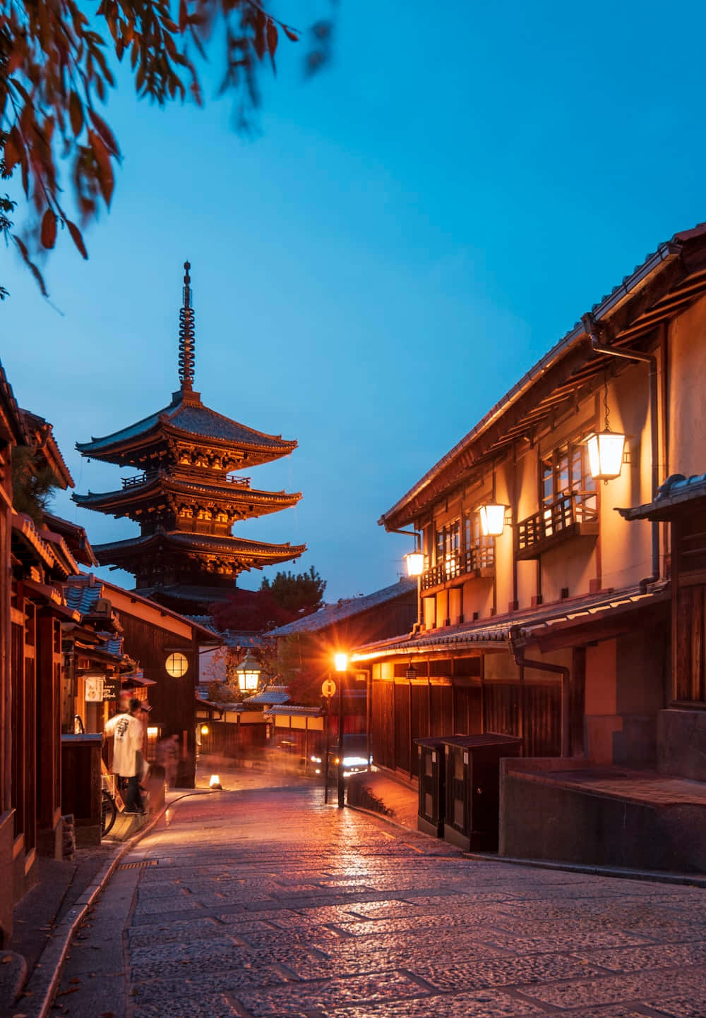 日本 桌布 壁紙 — 日本京都黃昏時分一條狹窄的鵝卵石街道，溫暖的燈籠在深藍色天空下閃耀，遠處矗立著一座寶塔。