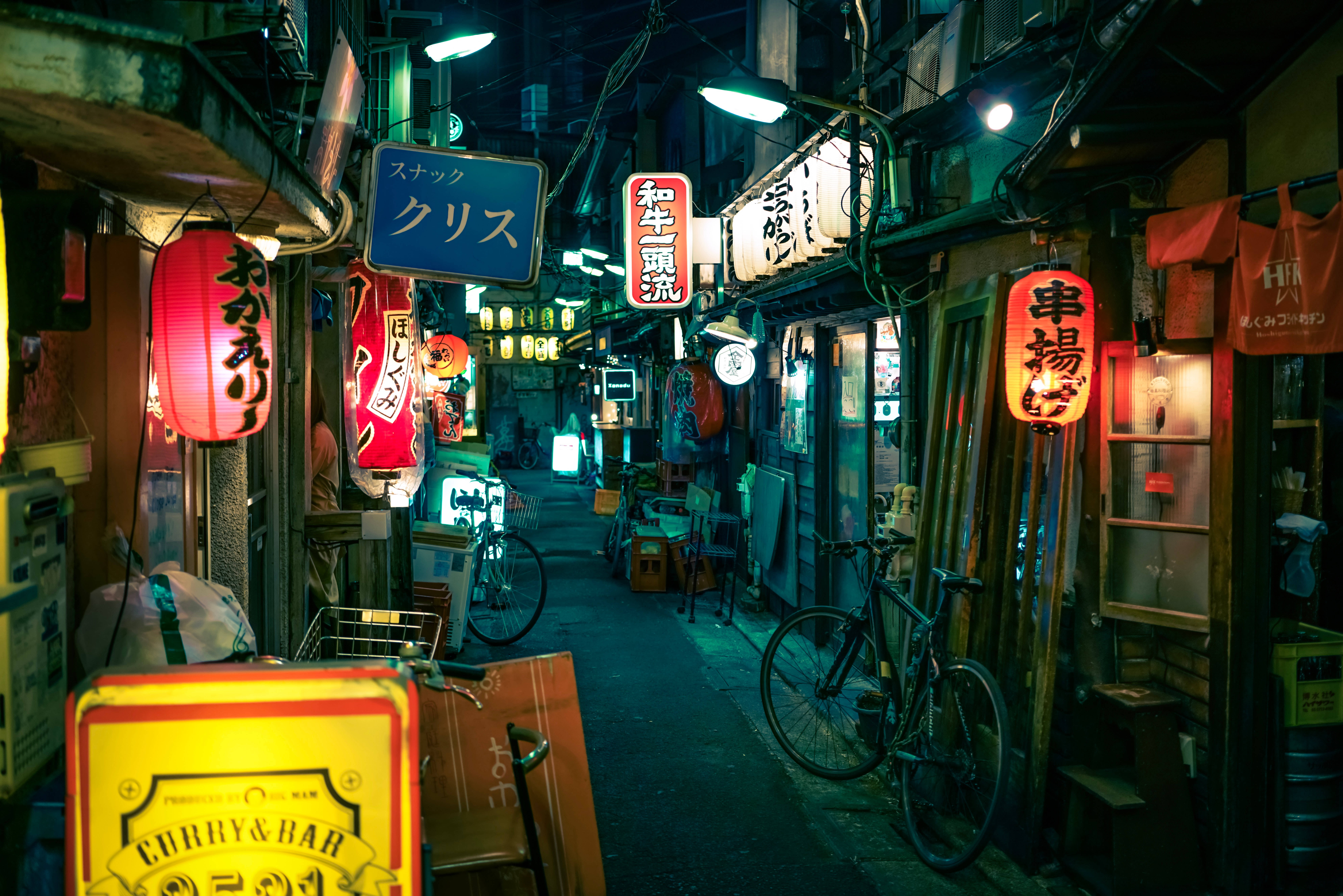 日本 桌布 壁紙 — 日本夜晚一條狹窄的霓虹燈小巷，有發光的招牌、腳踏車和木製店面，在潮濕的路面上投射出色彩繽紛的倒影。