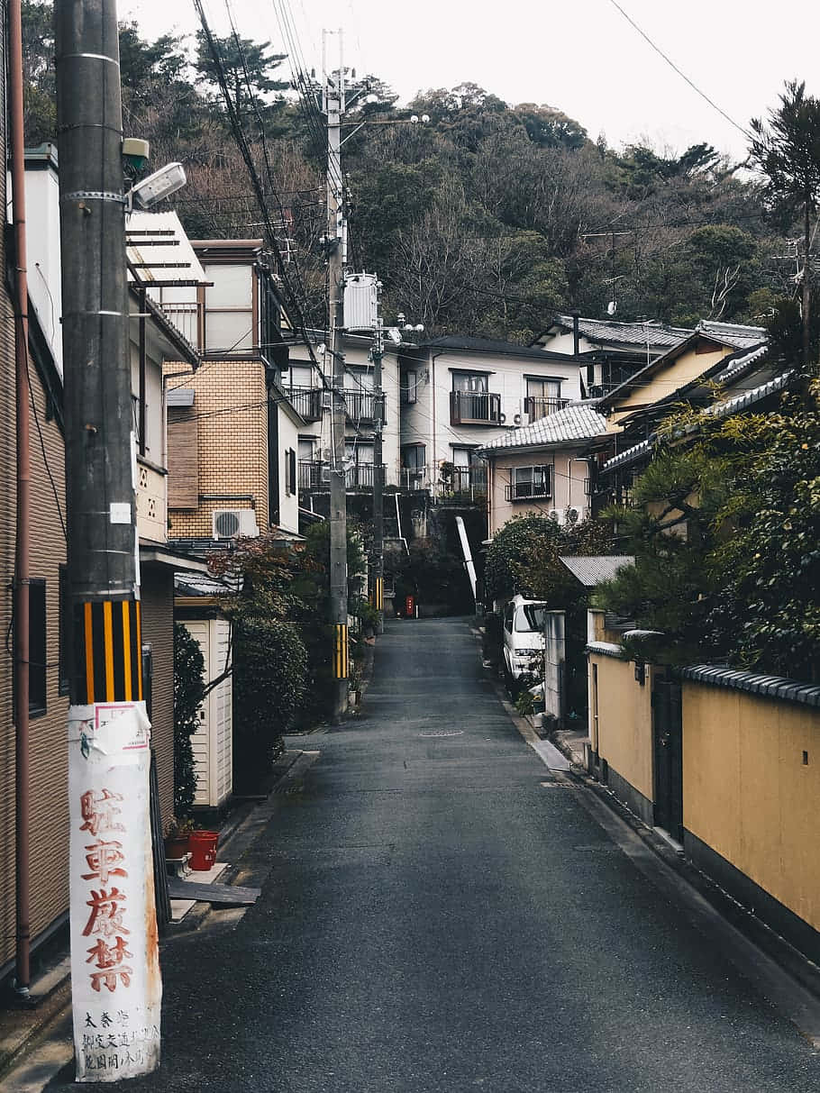 Neighborhood 桌布 壁紙 — 日本社區一條狹窄寧靜的街道，兩旁是傳統房屋和茂密的植被，天空多雲。