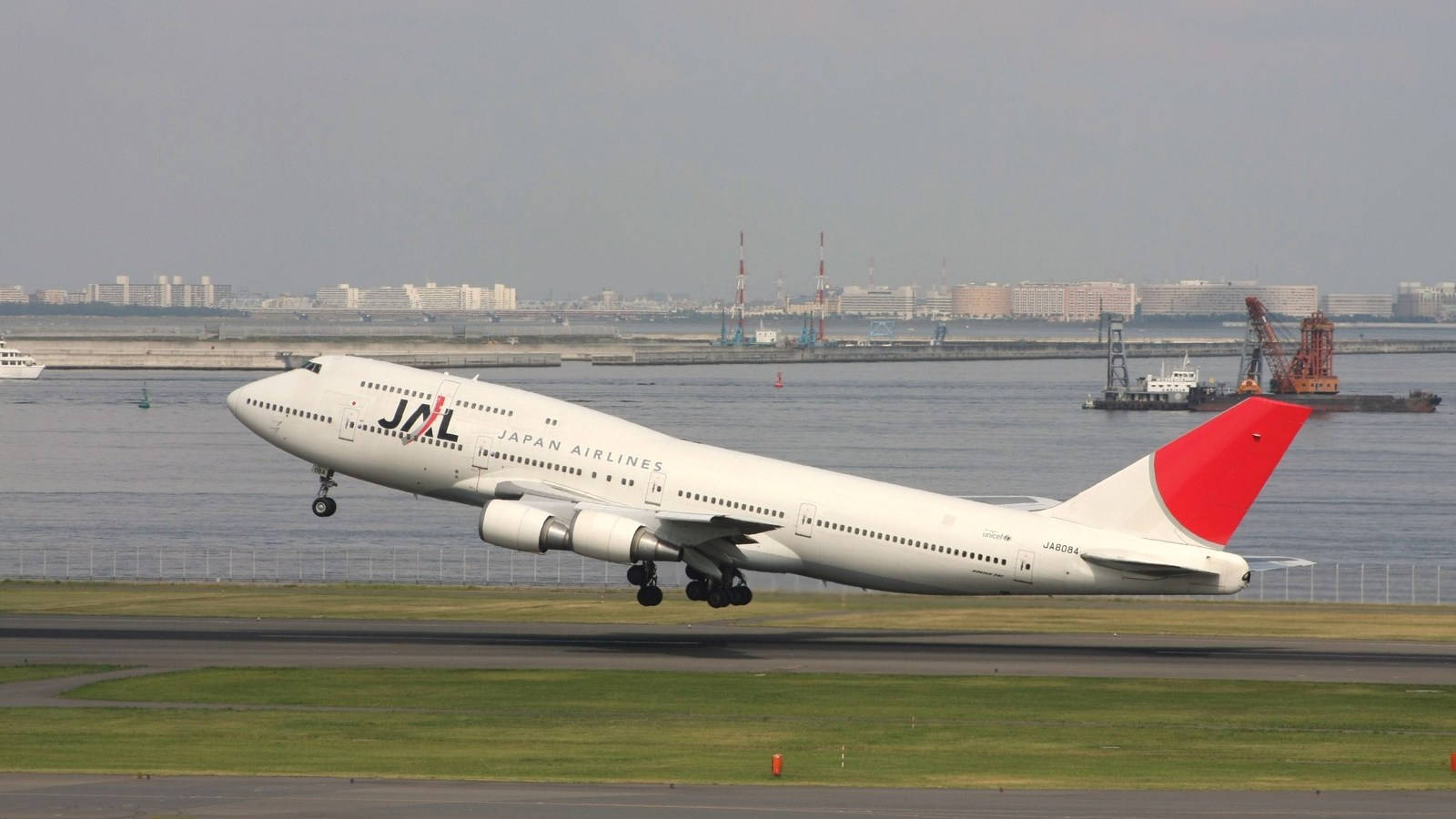 飛機 桌布 壁紙 — 日本航空的波音747在朦朧天空下，從海港旁的跑道起飛，遠處有城市建築。