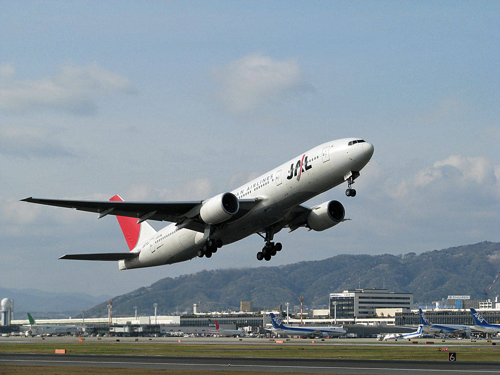 leaving 桌布 壁紙 — 日本航空的珍寶客機在部分多雲的天空下，從跑道起飛，背景有山脈和機場建築物。