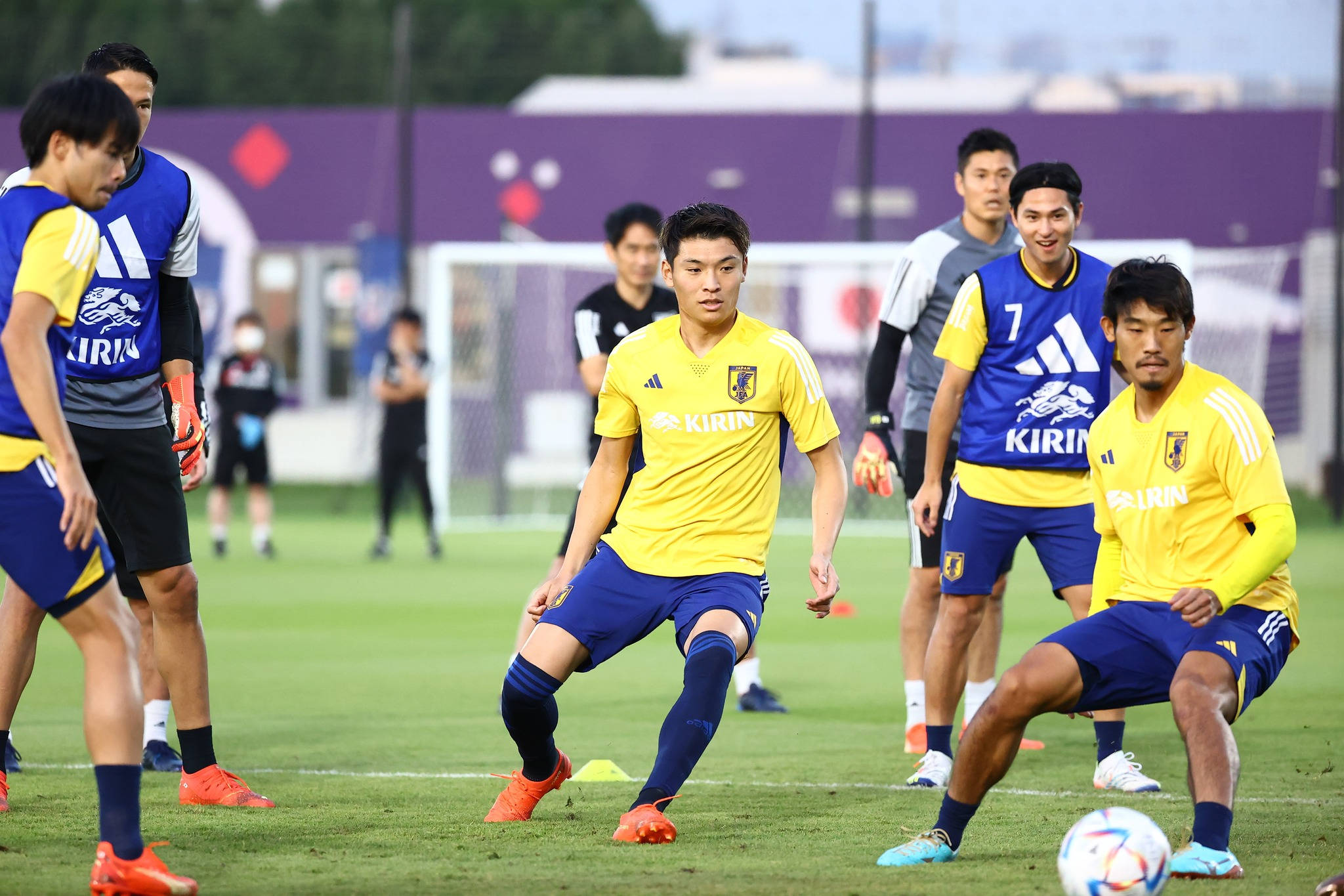 Football Players 桌布 壁紙 — 日本足球運動員身穿黃色和藍色球衣，在明亮的日光下於充滿活力的綠色球場上激烈訓練。