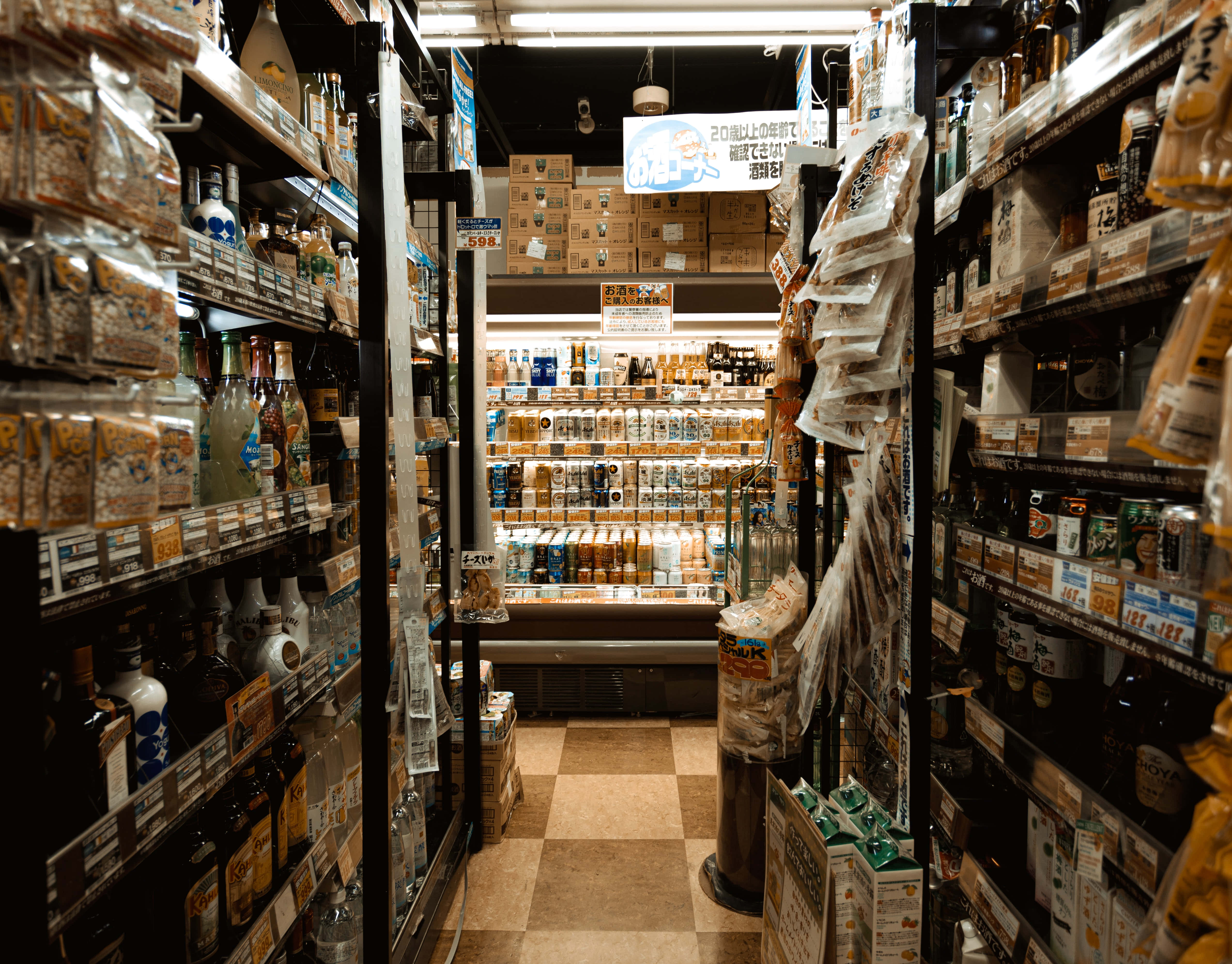 Grocery Store 桌布 壁紙 — 日本酒鋪內一條狹窄的通道，兩旁擺滿了瓶子和包裝商品的貨架，上方是明亮的螢光燈。