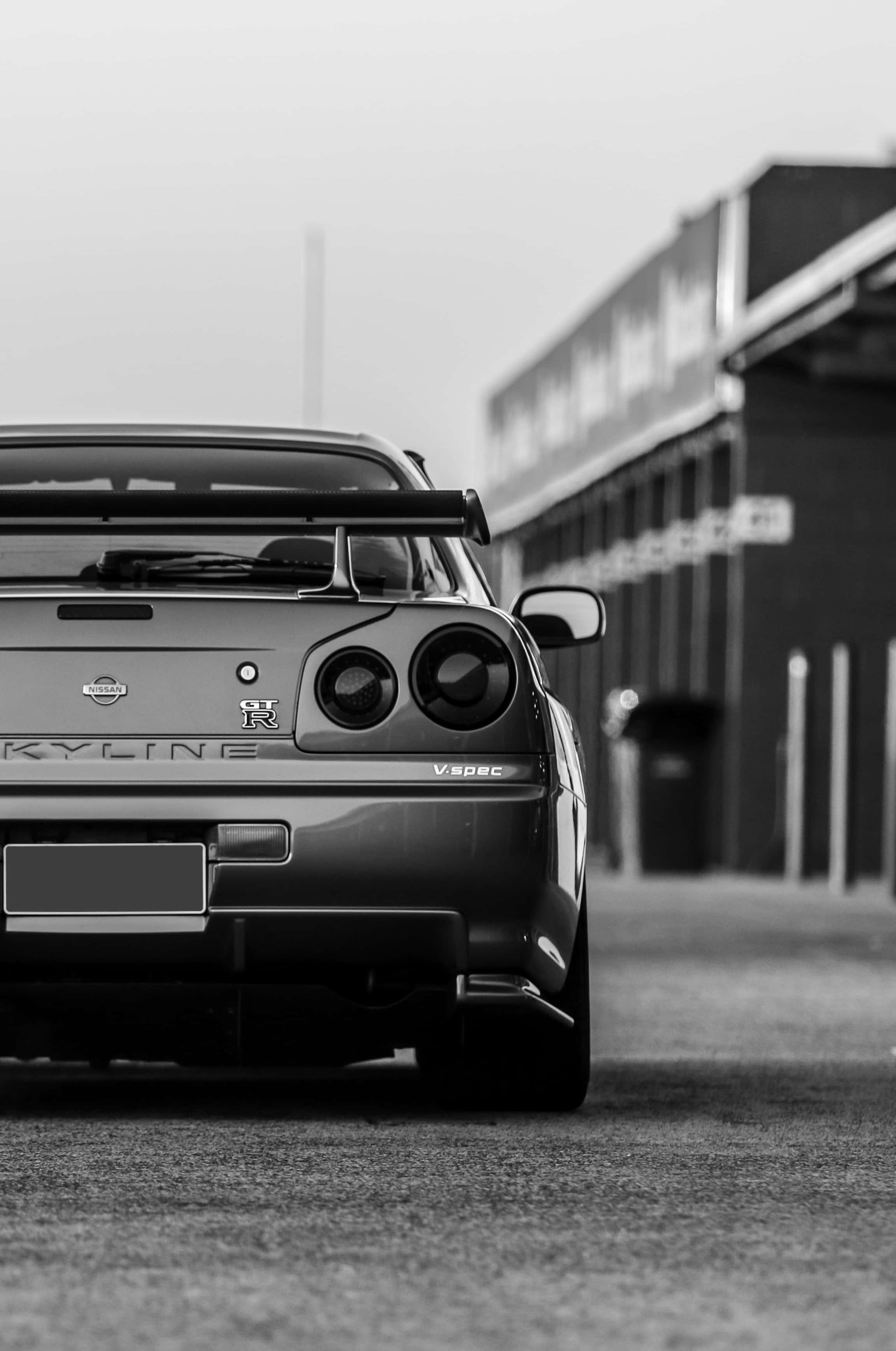 吉他 iPhone 桌布 壁紙 — 日產Skyline R34 GT-R停放在建築物前的黑白車尾視圖，背景模糊。