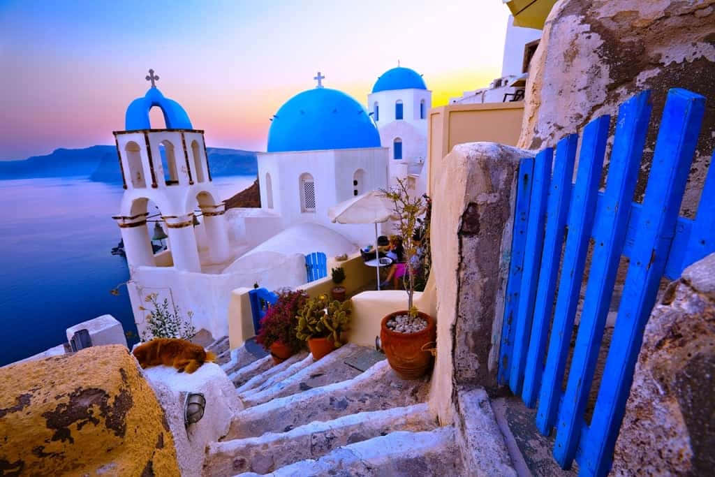Greek Island 桌布 壁紙 — 日落將聖托里尼標誌性的藍頂教堂和粉刷成白色的階梯沐浴在溫暖的金色光芒中。